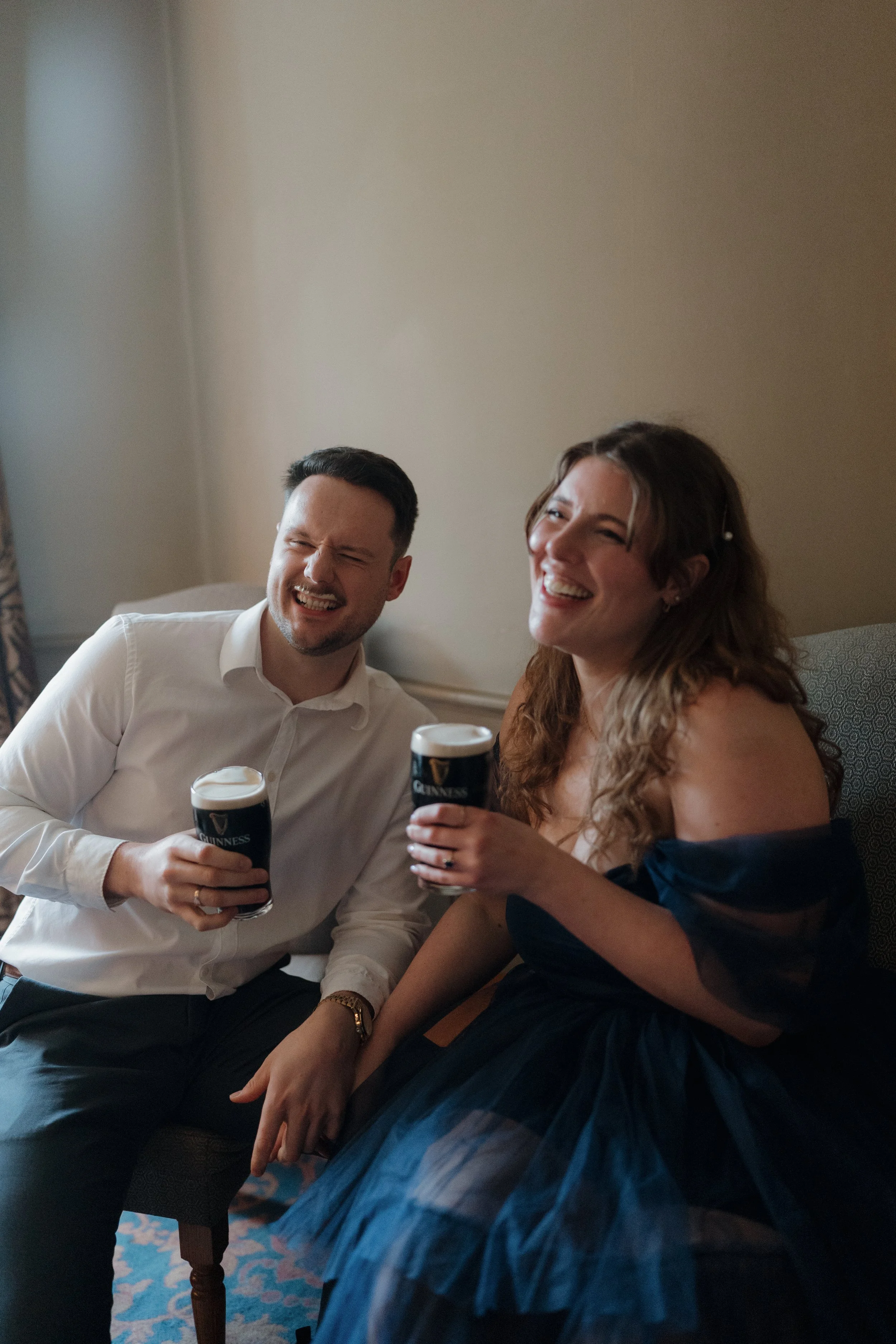 A man and woman sitting on a couch, smiling and holding glasses of Guinness beer, enjoying a moment together.