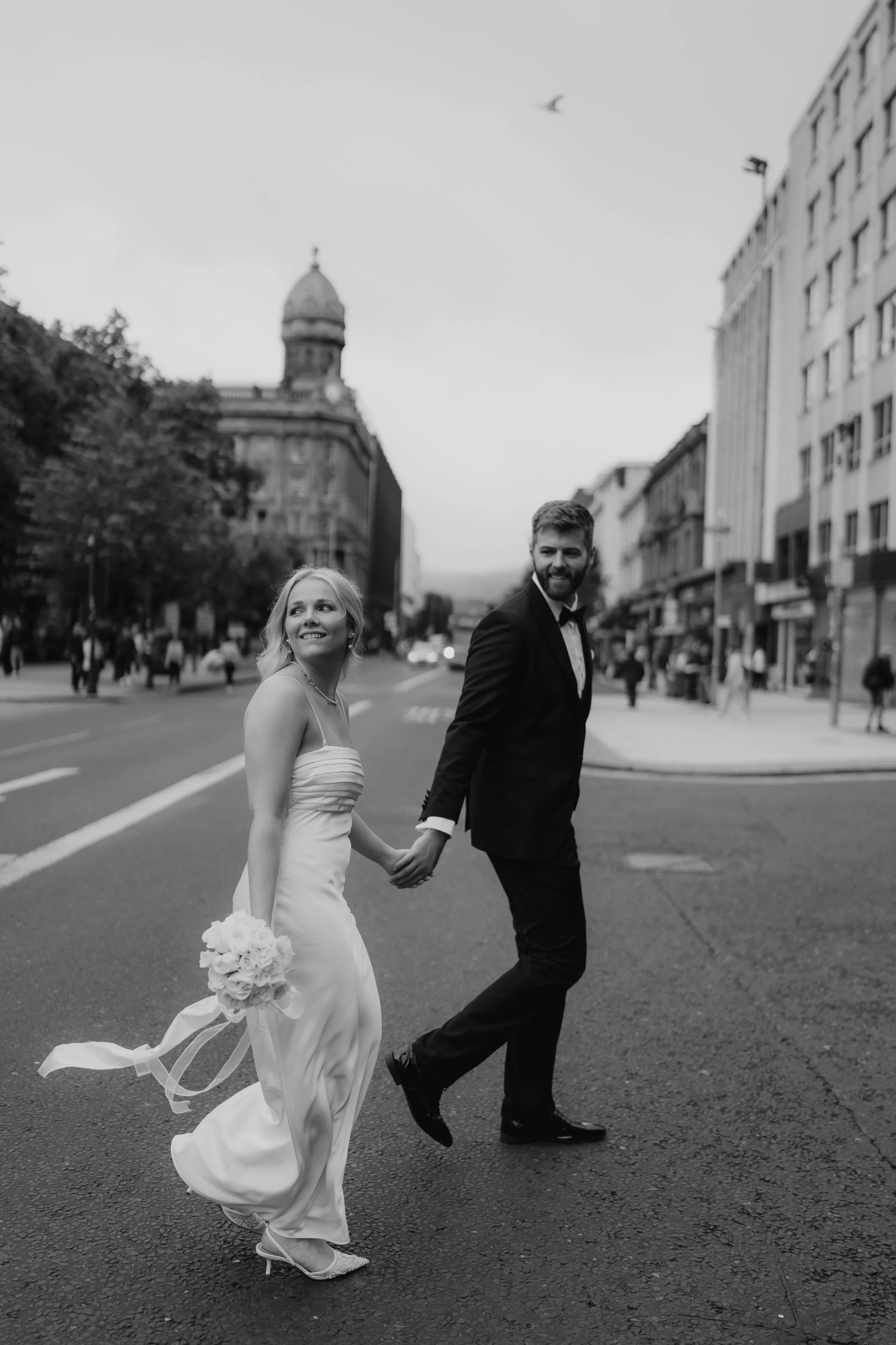 Black and white photo of a newlywed couple holding hands and crossing a city street. The woman is in a wedding dress holding a bouquet, and the man is in a tuxedo. They are smiling and looking at each other.