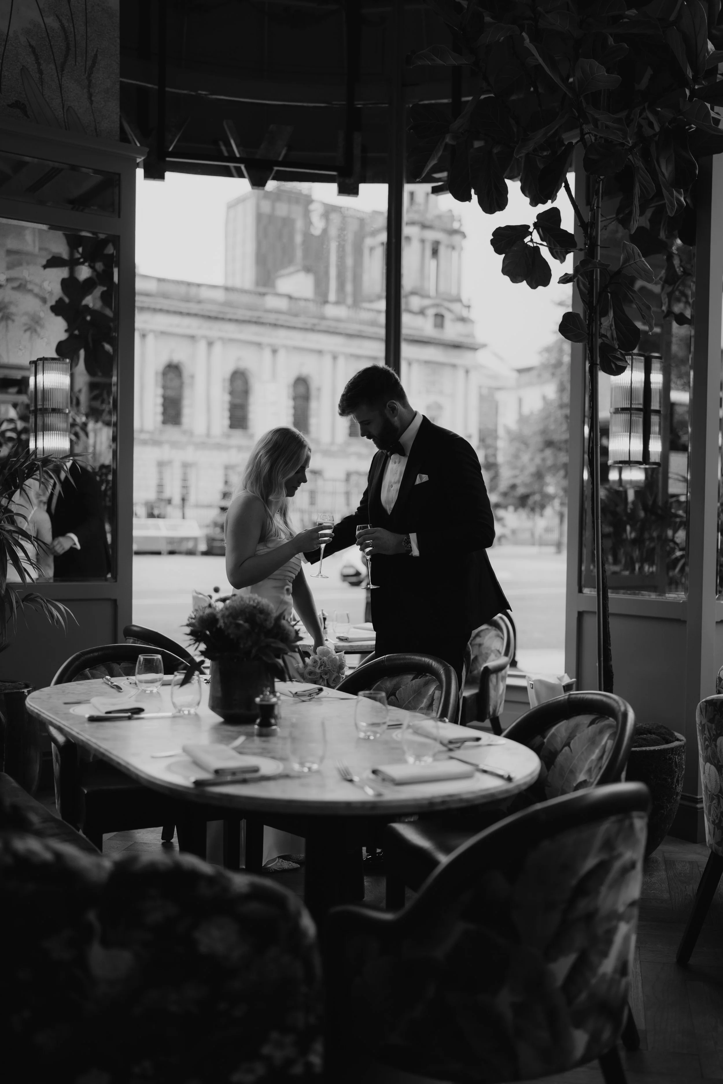 Black and white photo of a couple in wedding attire standing inside a restaurant, holding glasses of wine, with large windows showing a cityscape in the background at The Ivy, Belfast.