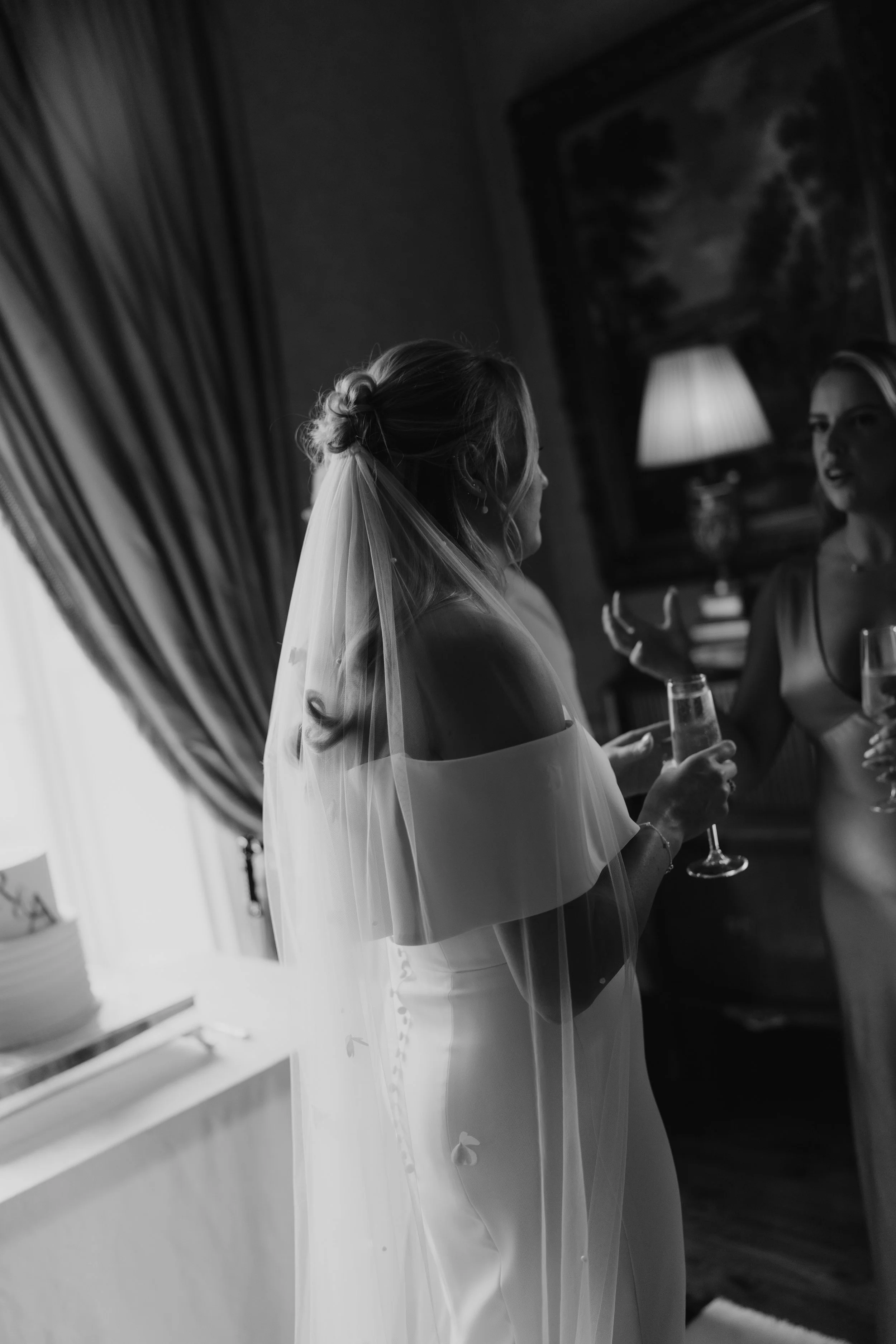 Bridal woman in wedding dress and veil holding a glass of champagne, talking to a woman in a sleeveless dress, in a dimly lit room with curtains and a lamp in the background.