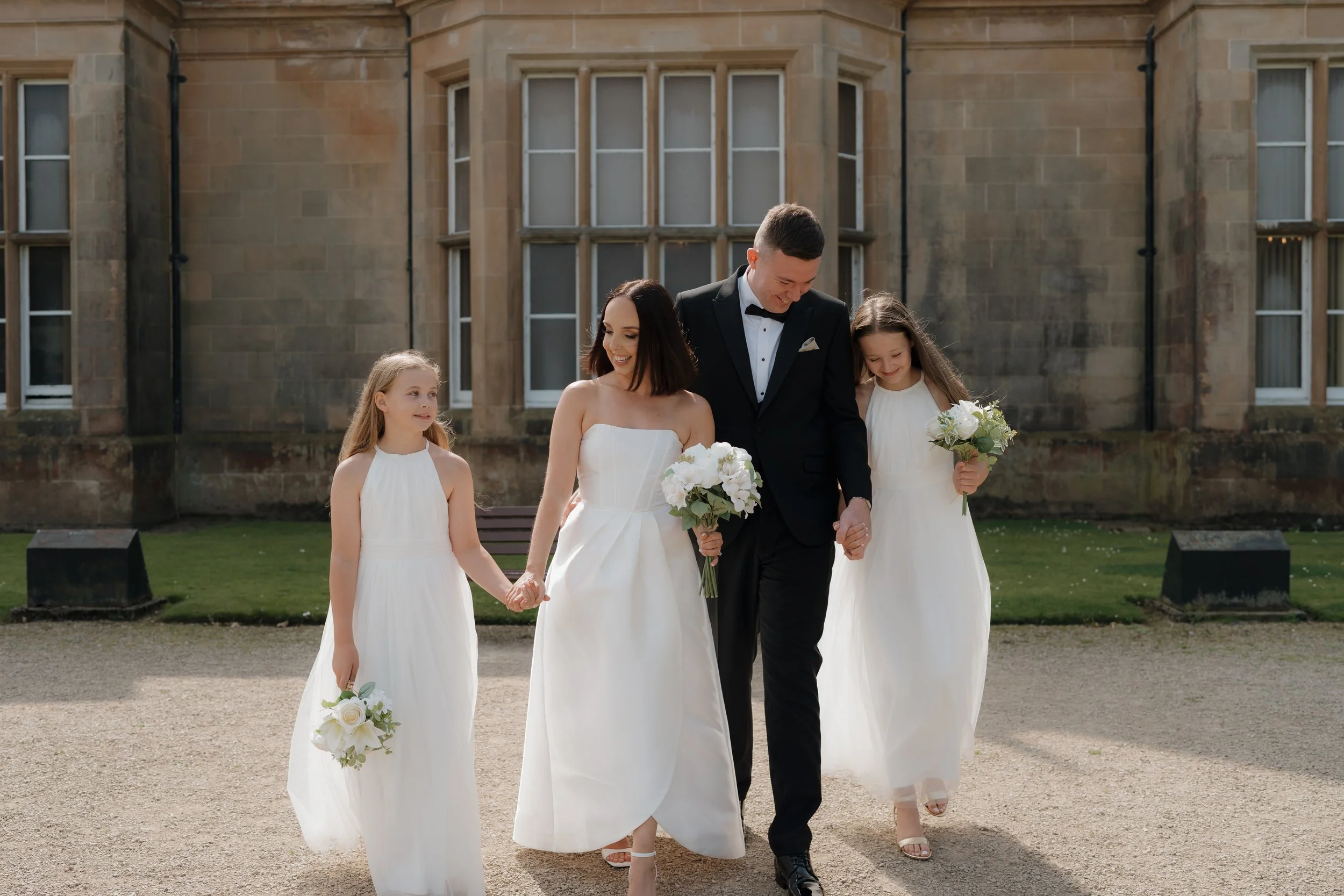 Bride and groom walking outside with two young girls, all holding bouquets, in front of a historic stone building, Bangor Castle.