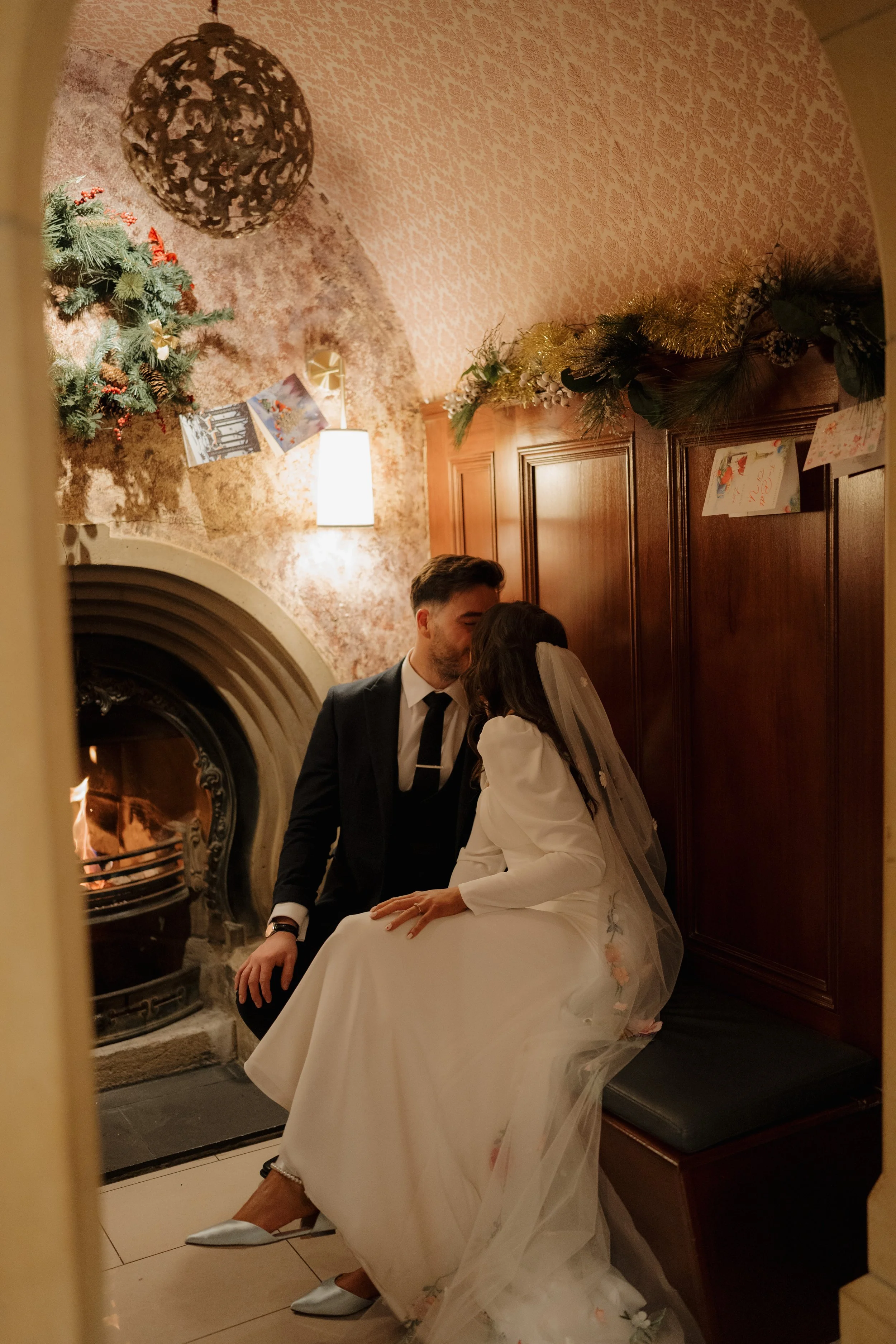 A couple dressed in wedding attire sharing an intimate moment by a fireplace decorated with Christmas garland and cards, inside a warmly lit room.