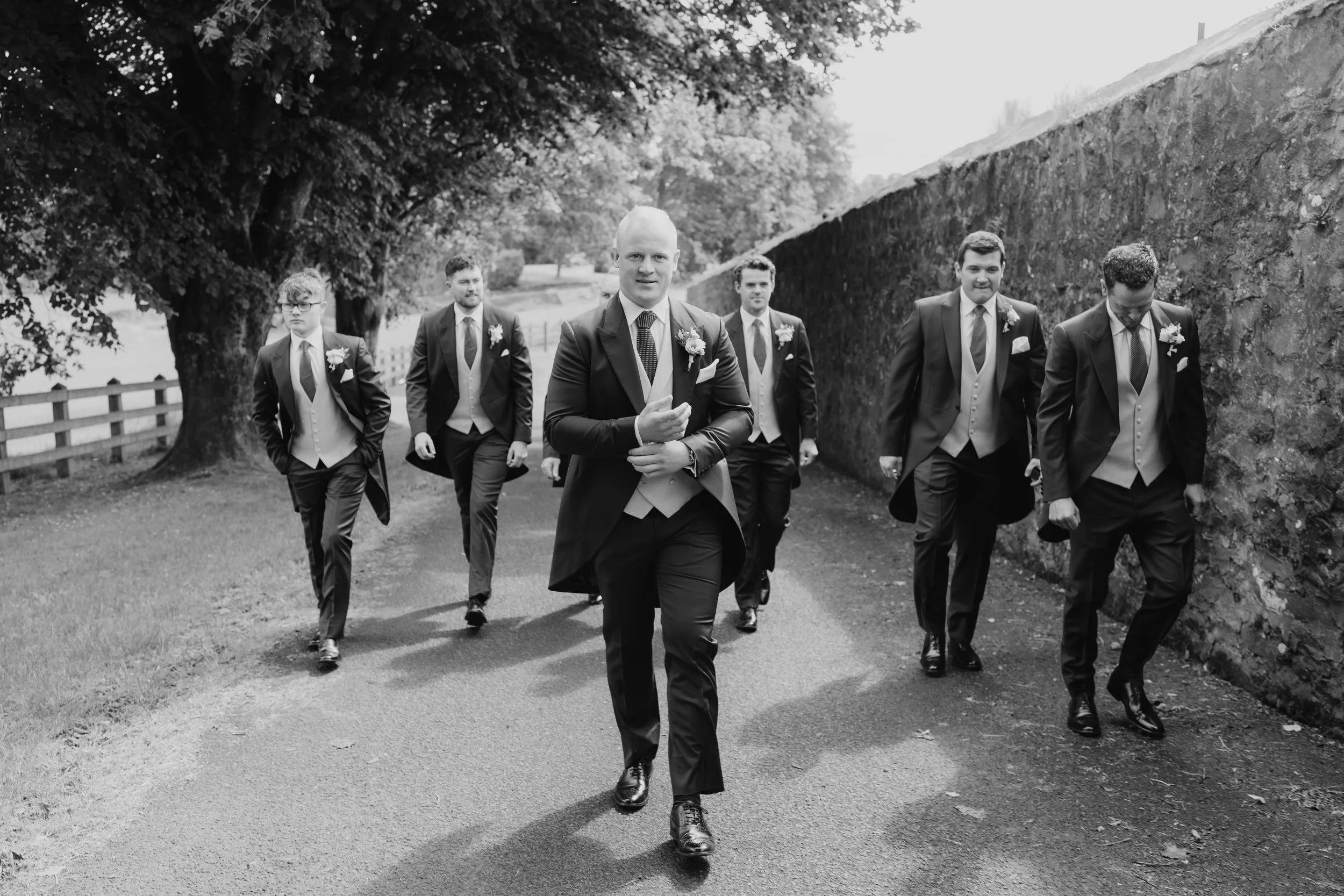 A group of seven men dressed in suits and ties, walking outdoors, with one man in the forefront, potentially a groom with six groomsmen, in a scenic outdoor setting with trees and a hedge in the background.
