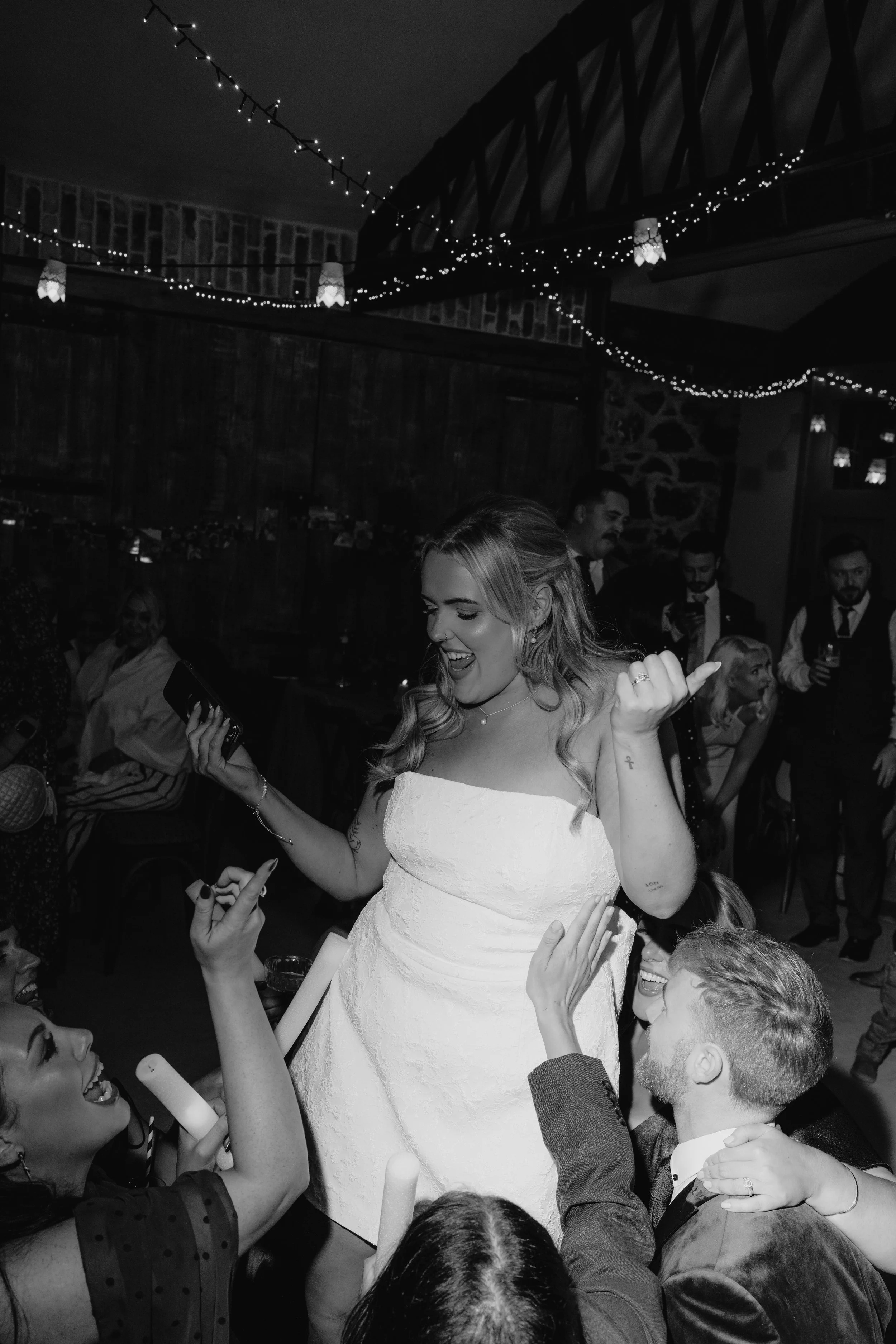 A black-and-white photo of a woman in a wedding dress dancing and celebrating with friends at a wedding reception, with string lights hanging overhead.