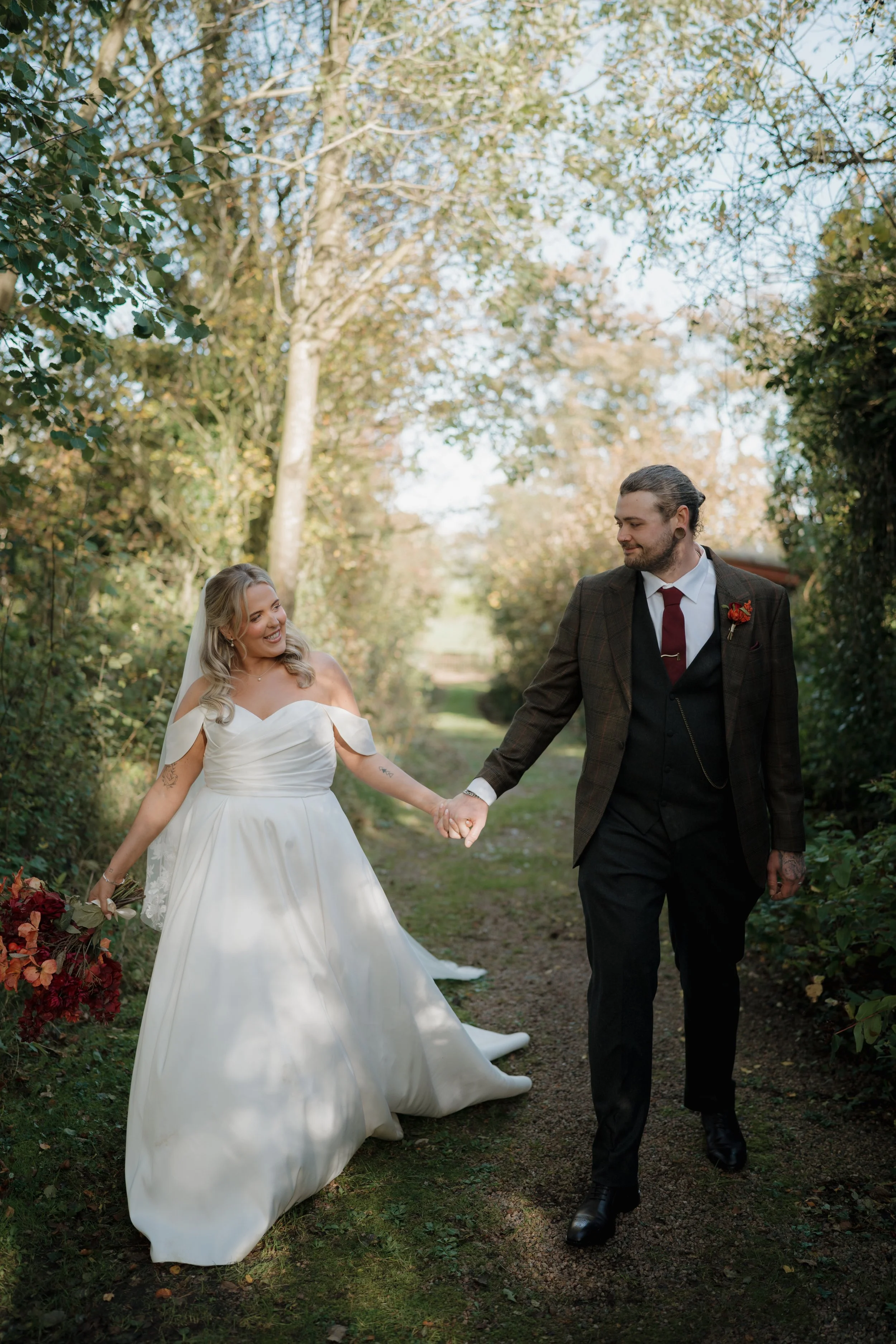 A bride and groom holding hands and walking in a wooded outdoor setting, smiling at each other.