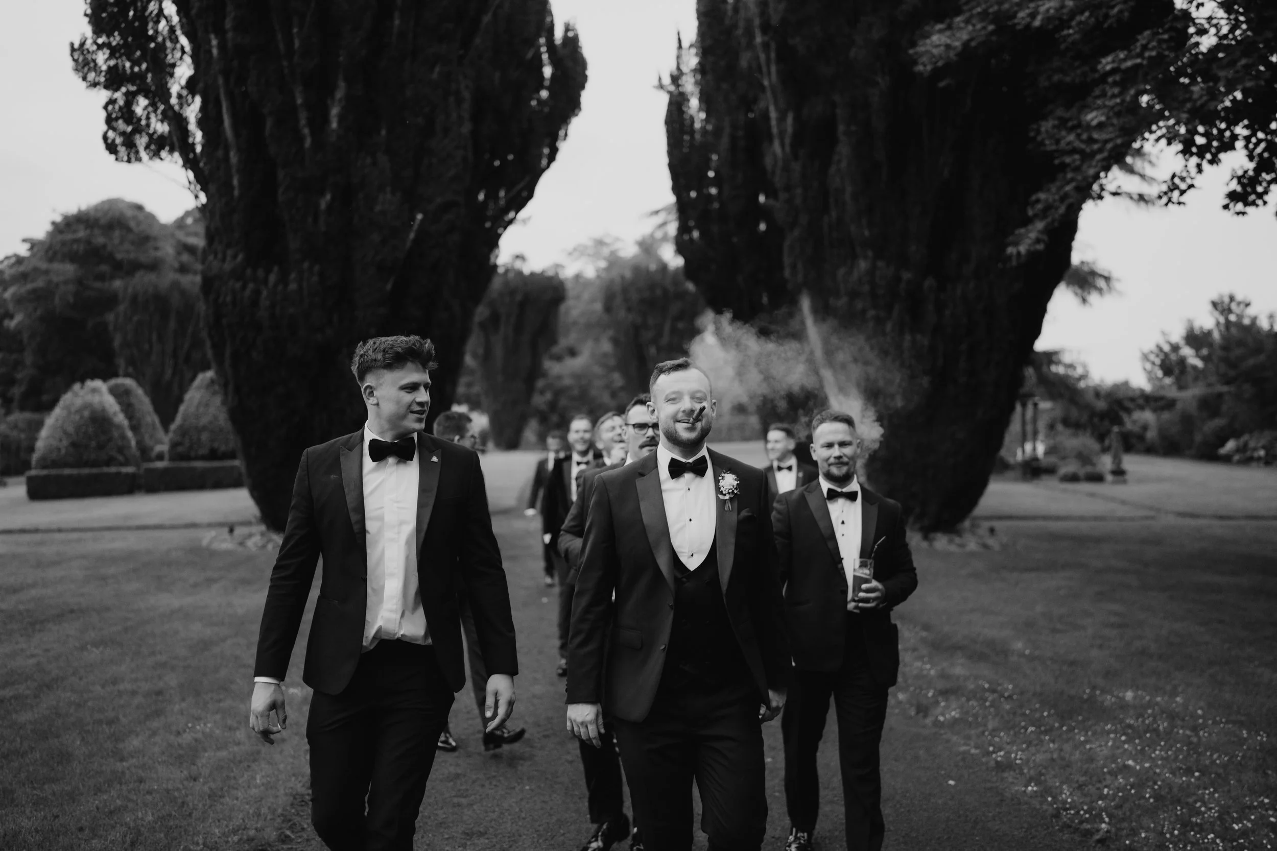 A group of men in tuxedos walking outdoors on a grassy area with large trees in the background, smiling and carrying drinks at Tankardstown House.