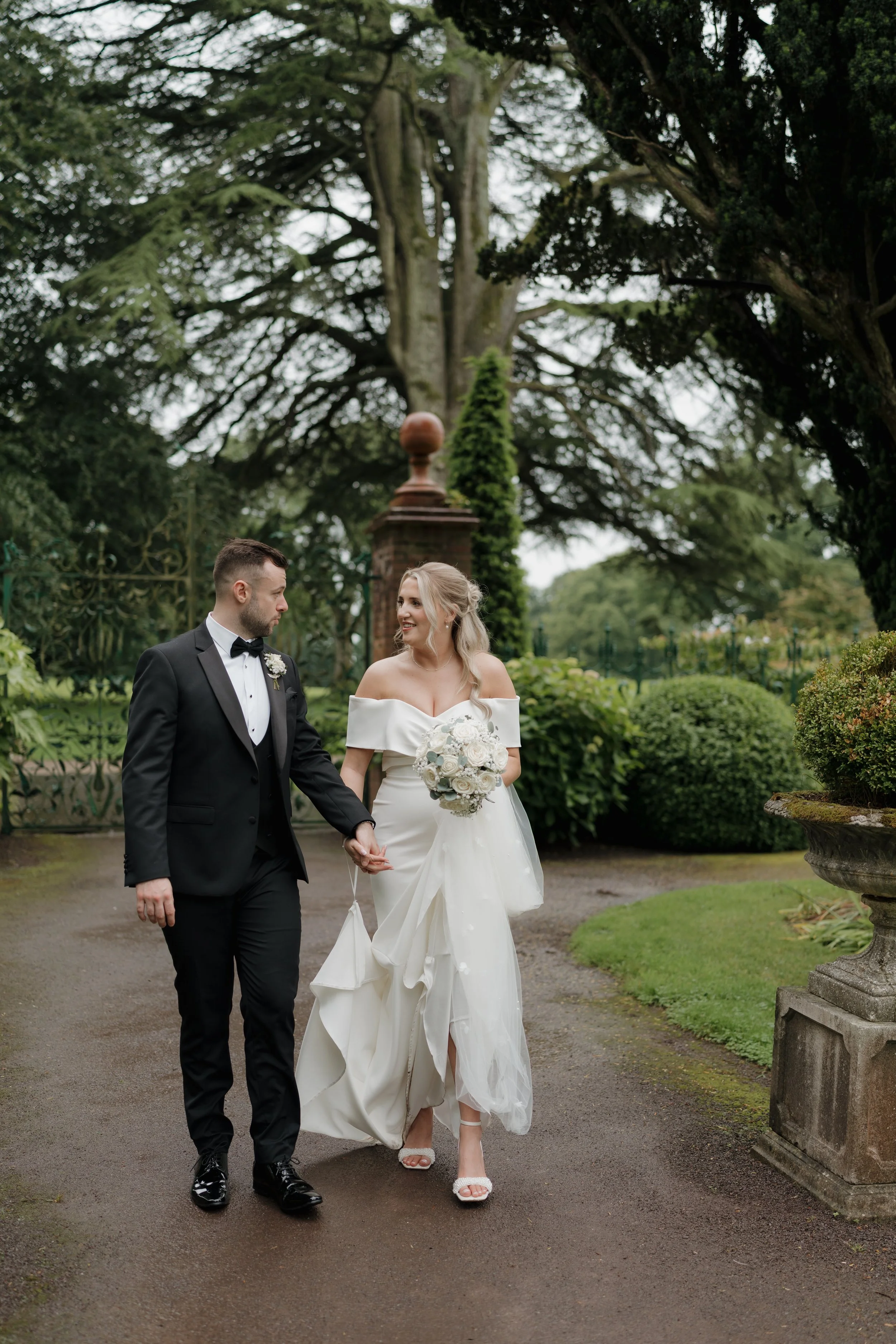 Bride and groom walking outdoors in a garden, holding hands, with the bride holding a bouquet of white flowers.