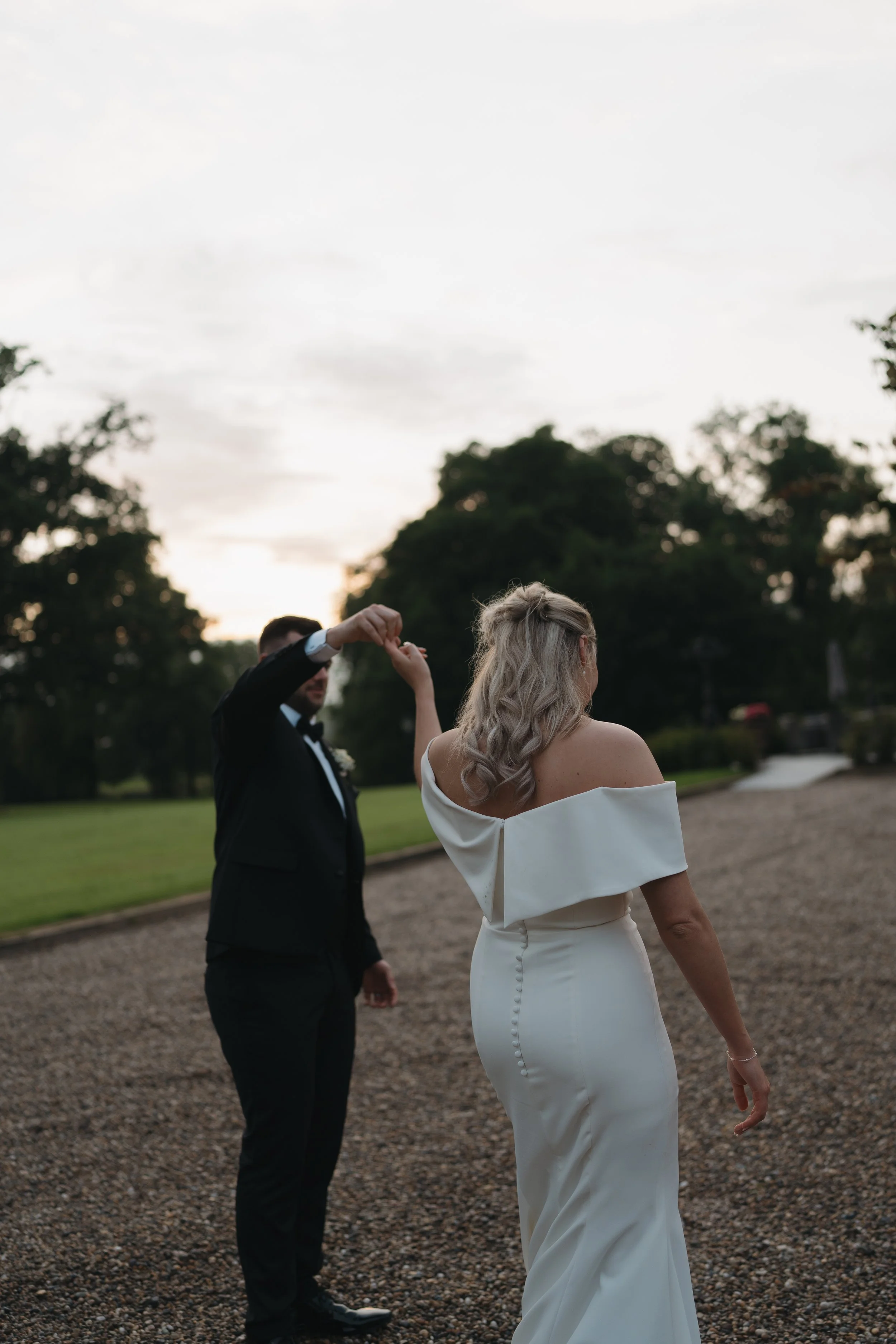 A bride and groom dancing outdoors during sunset, with trees and a gravel path in the background.