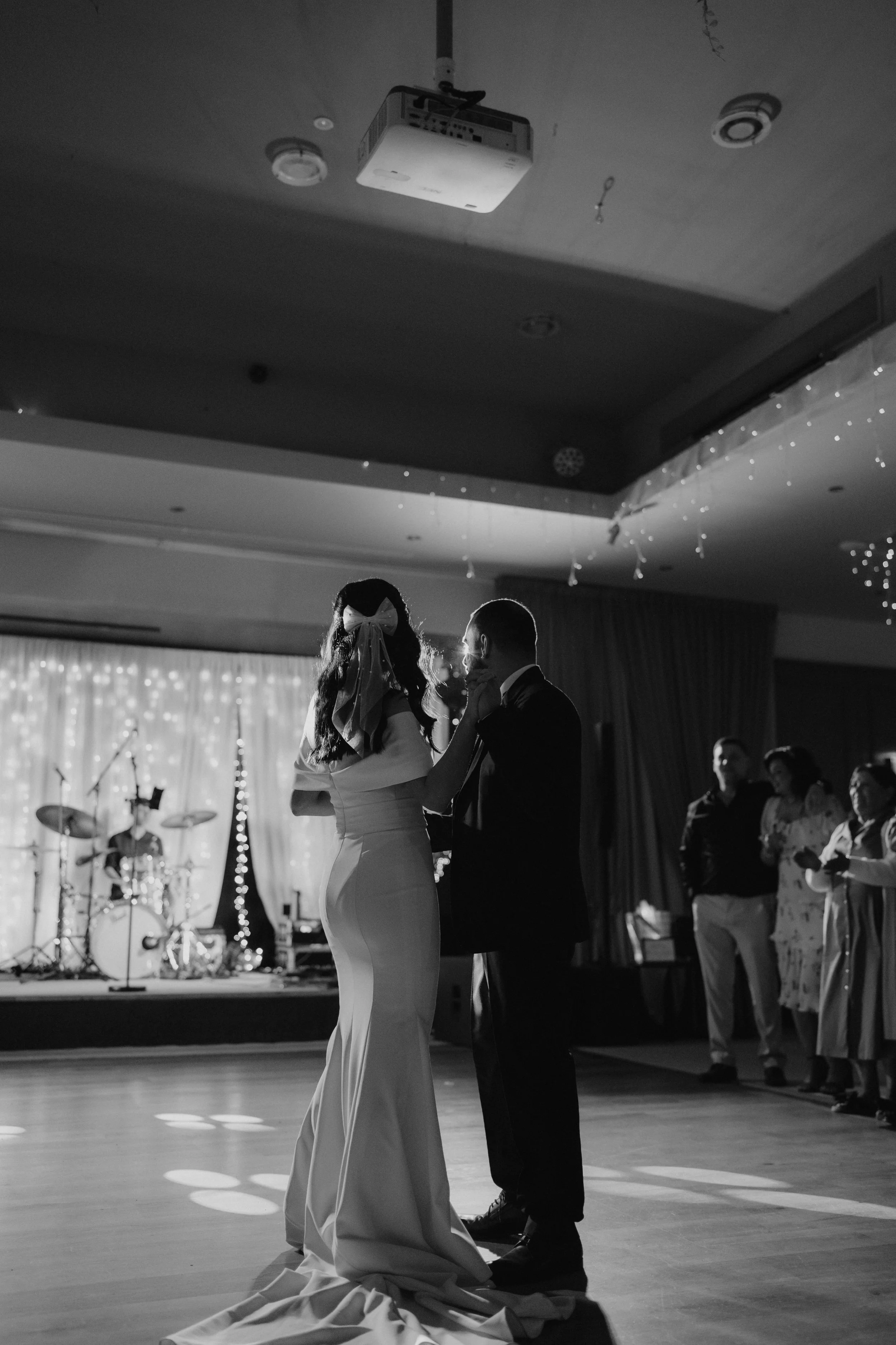 A black and white photo of a bride and groom dancing at their wedding reception, with guests watching in the background and a band setup visible on a stage.