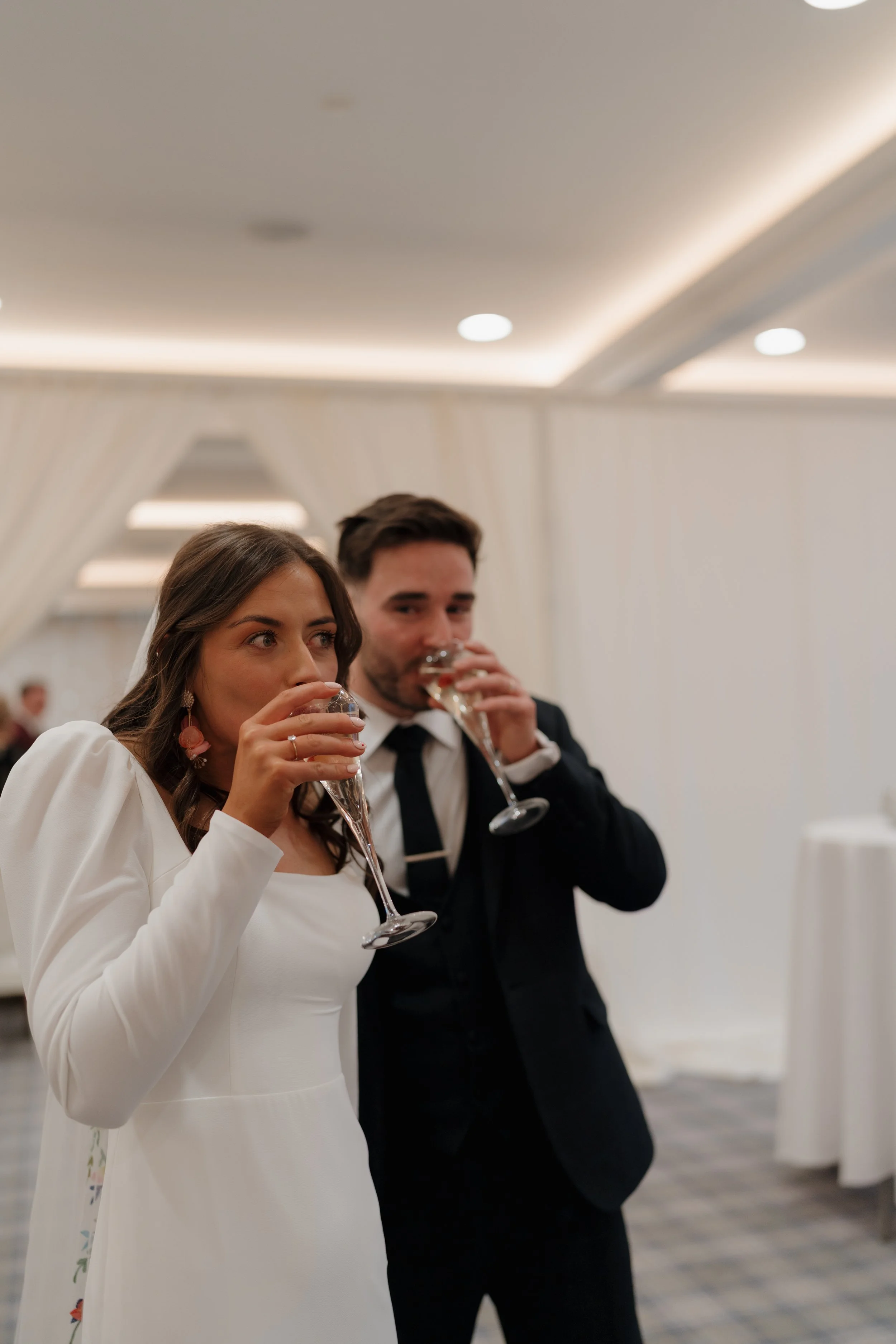 A woman in a white dress and a man in a black suit and tie are drinking champagne at a formal event, likely a wedding reception, in an elegant indoor setting.