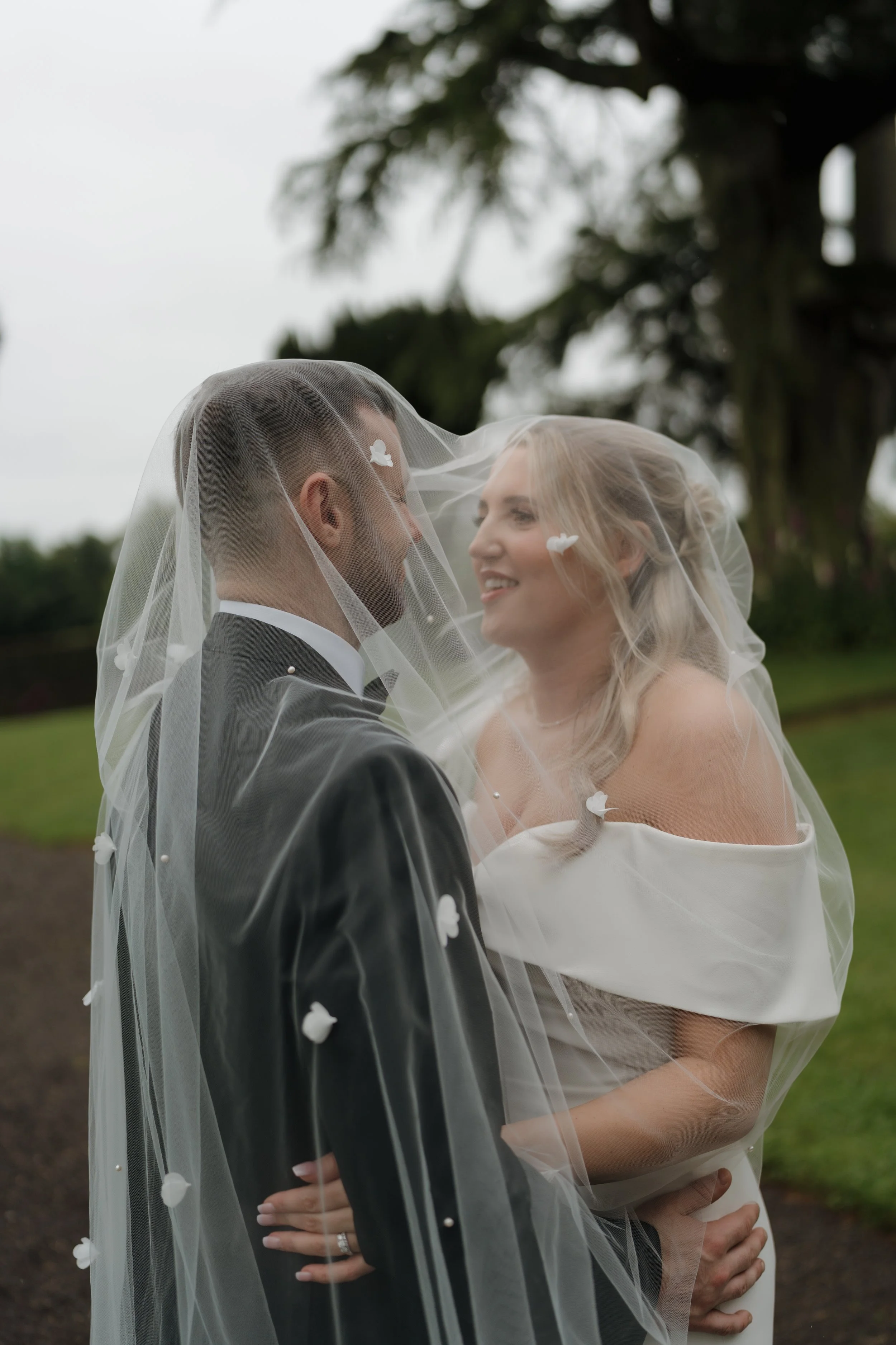 A bride and groom gently touching noses under a veil decorated with small flowers outdoors.
