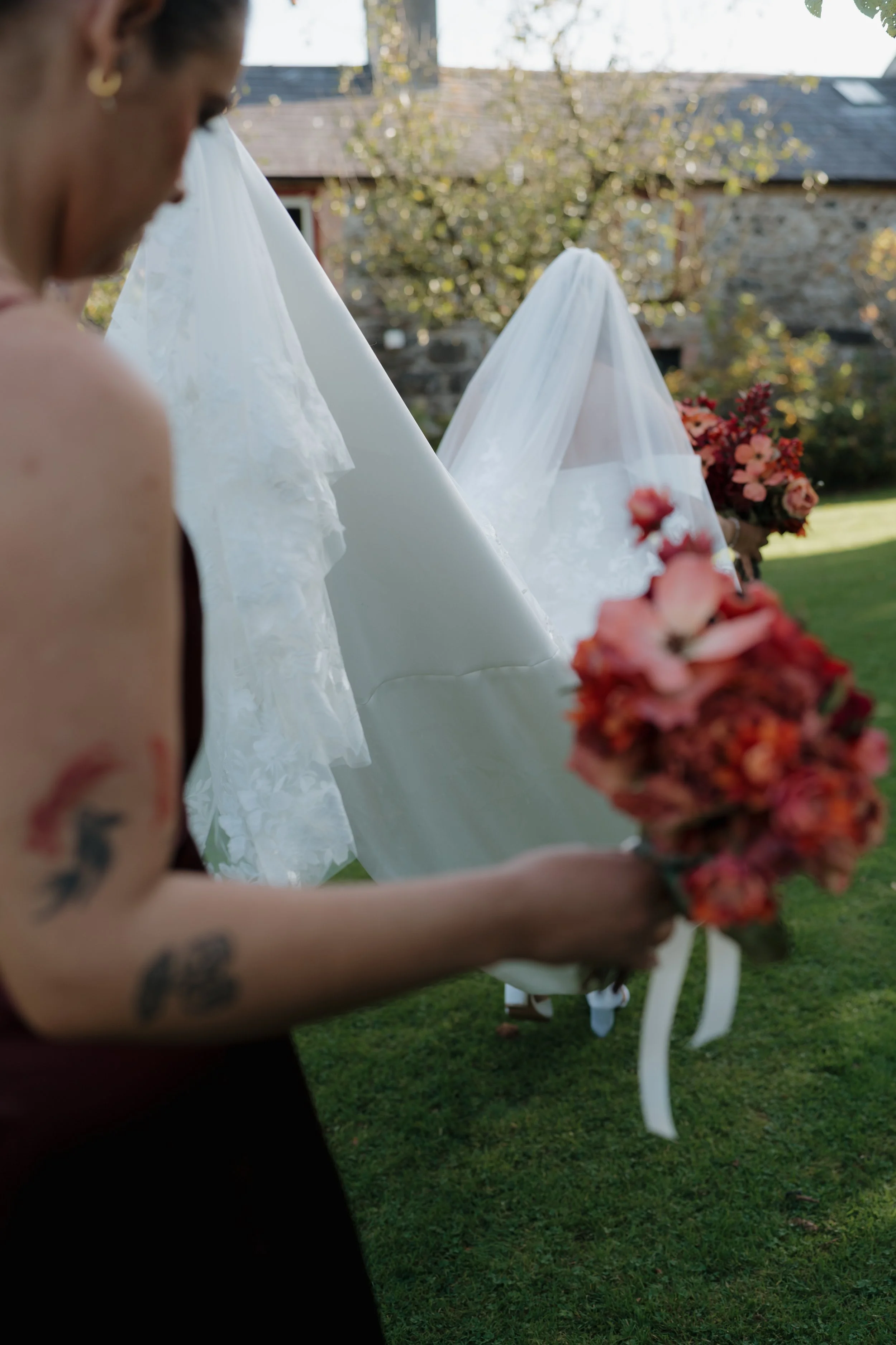 A woman with tattoos on her arm holding a bouquet of red and pink flowers outdoors, with tents and a stone building in the background.