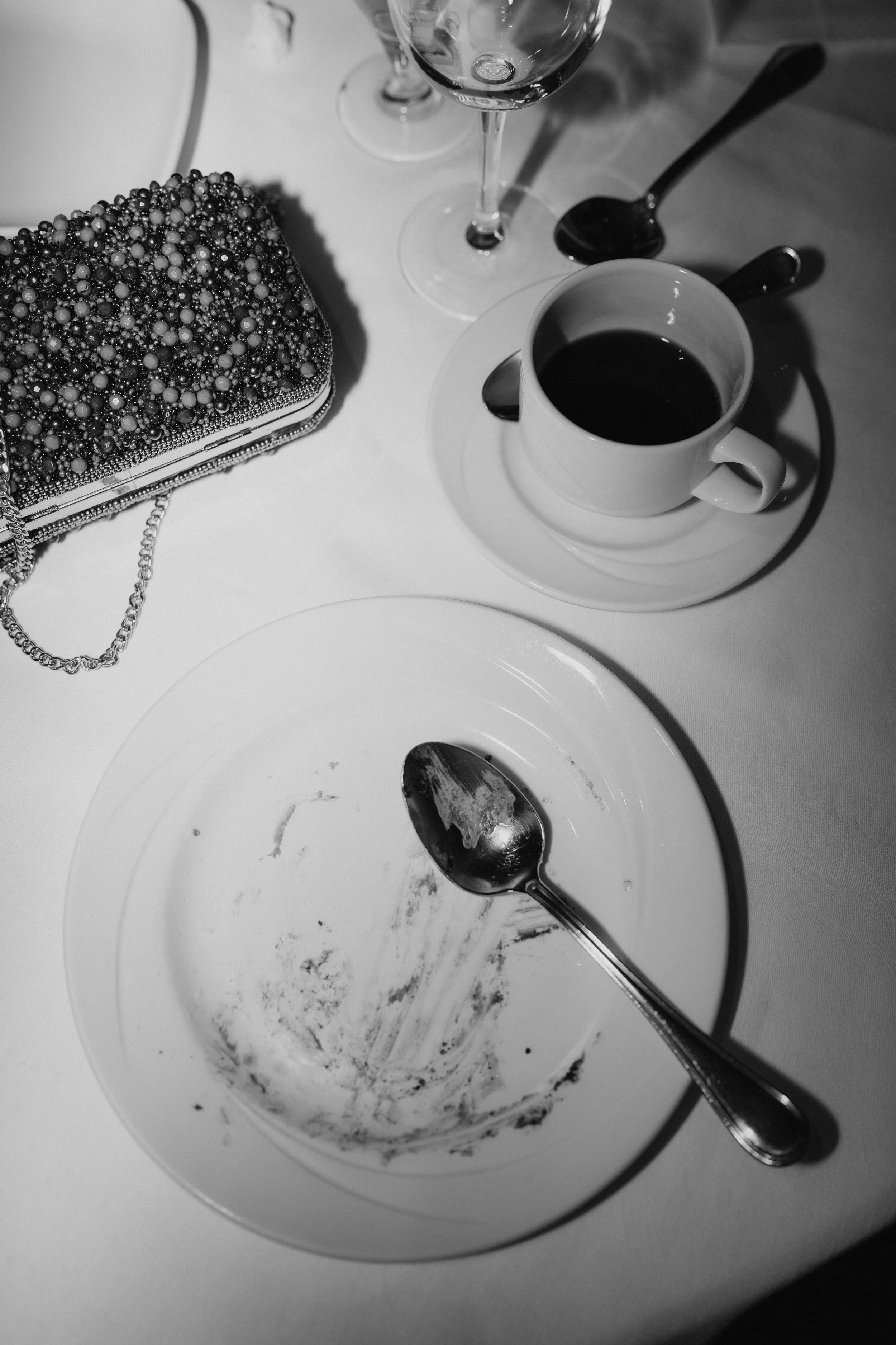 Empty plate with a spoon, a cup of coffee on a saucer, two glasses, a beaded clutch, and a spoon with some leftover food on a white table.