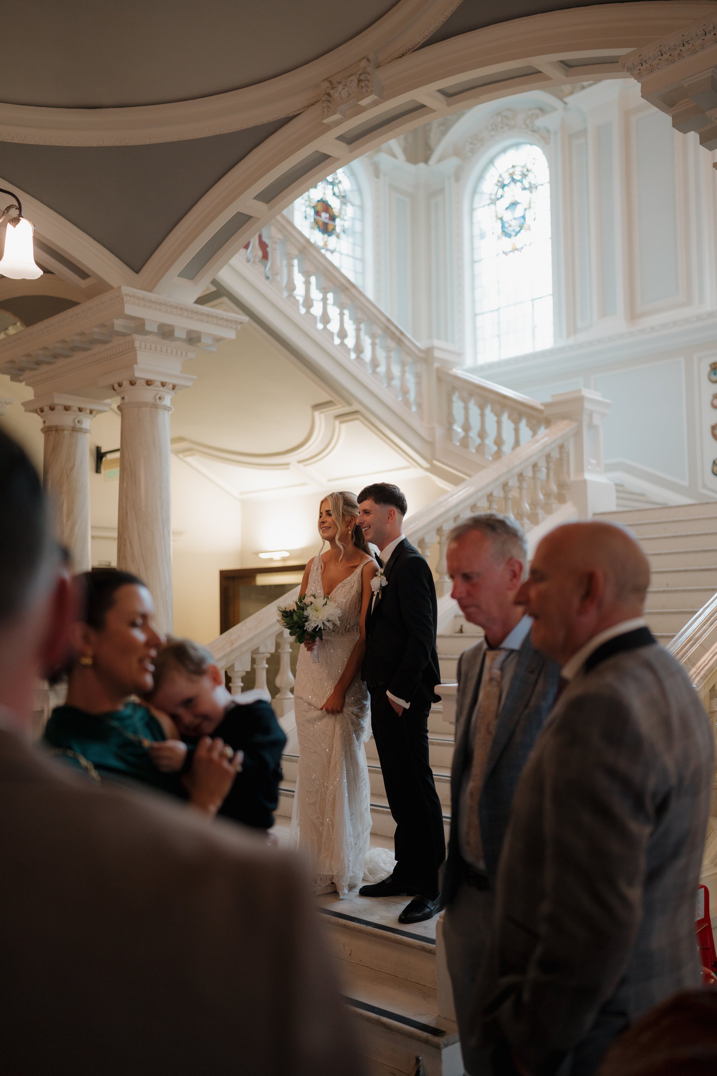 A bride and groom standing on a staircase during their wedding ceremony, surrounded by guests inside a grand, elegant church with stained glass windows.
