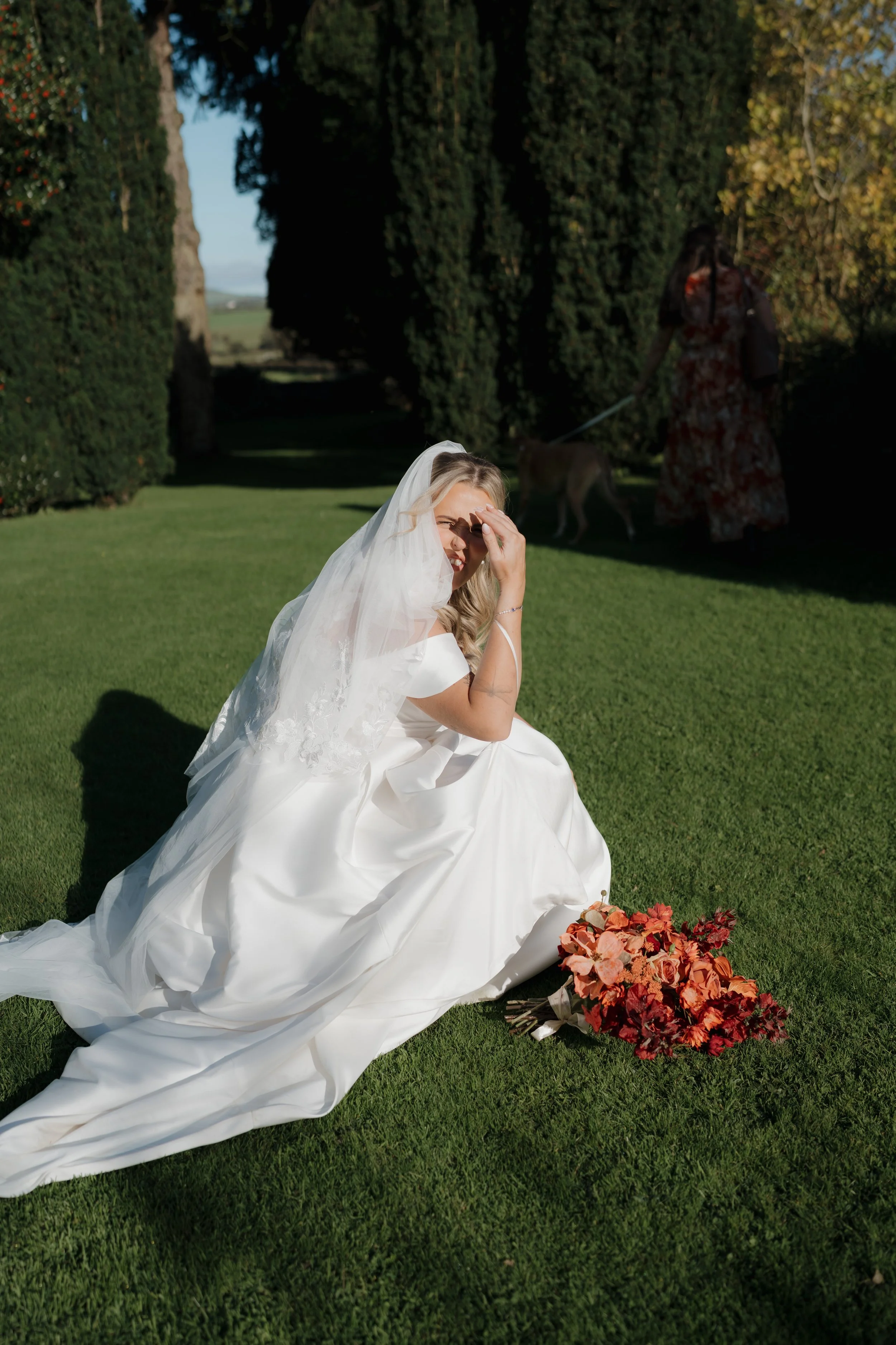 Bride sitting on green grass in wedding dress with veil, smiling and shading her eyes, with a bouquet of red and peach flowers nearby, and a woman walking a dog in the background.