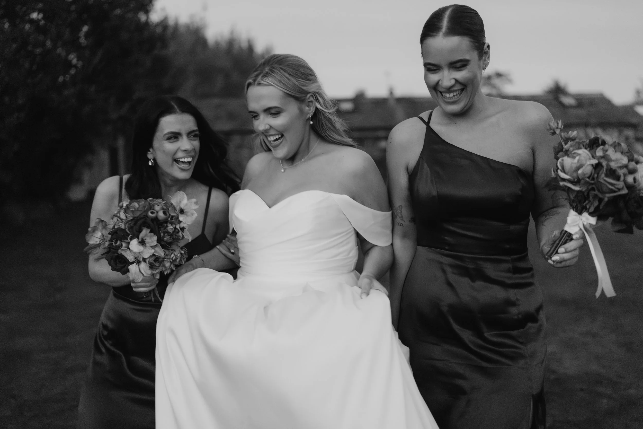 Black and white photo of three women, wedding and bridesmaids, walking outdoors, laughing, with bouquets of flowers.