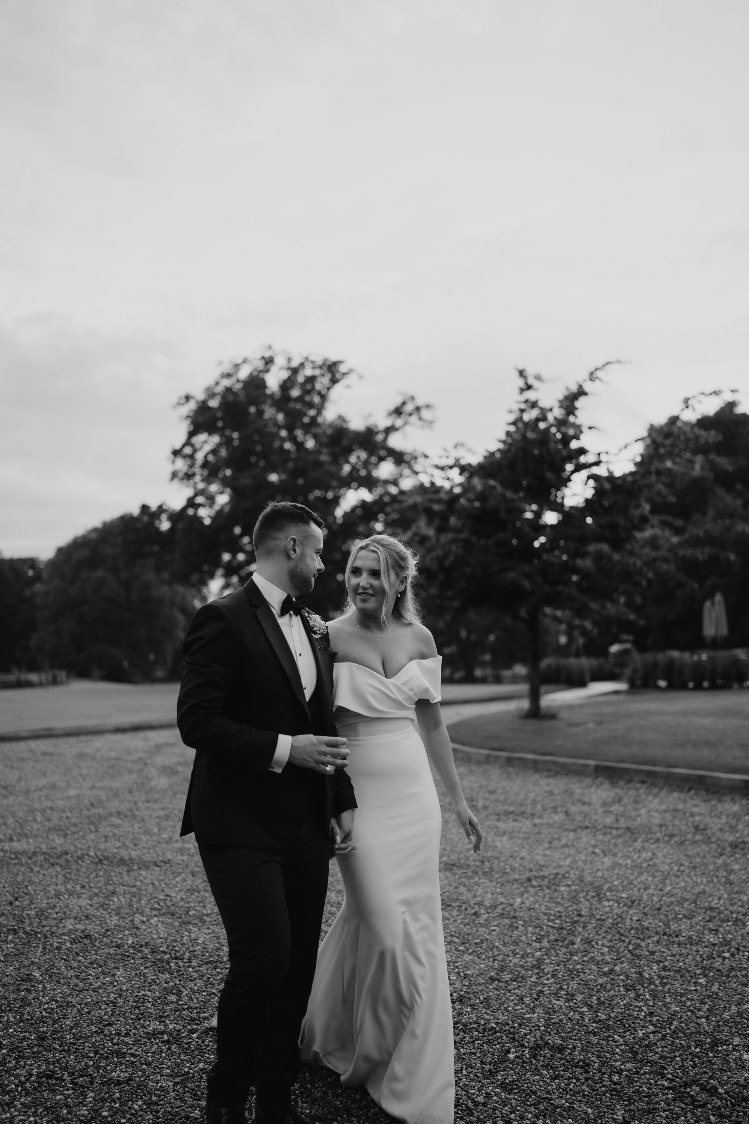 Black and white photo of a bride and groom walking outdoors on a gravel path, surrounded by trees, during a wedding celebration.