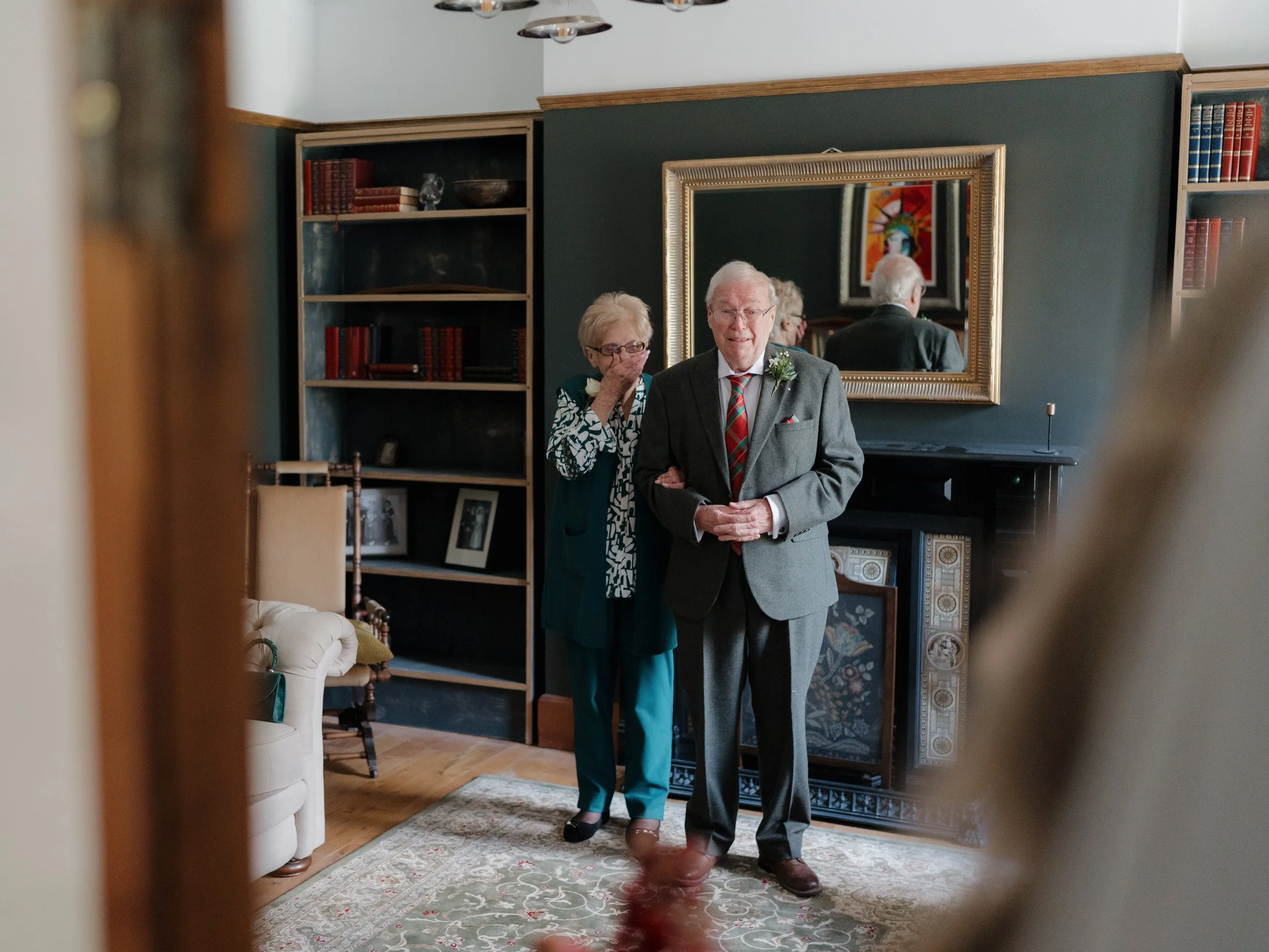 An elderly couple stands together in a living room, with the man smiling and the woman covering her mouth. The room has a dark green wall, a large mirror, a black fireplace, and shelves with books and photo frames.
