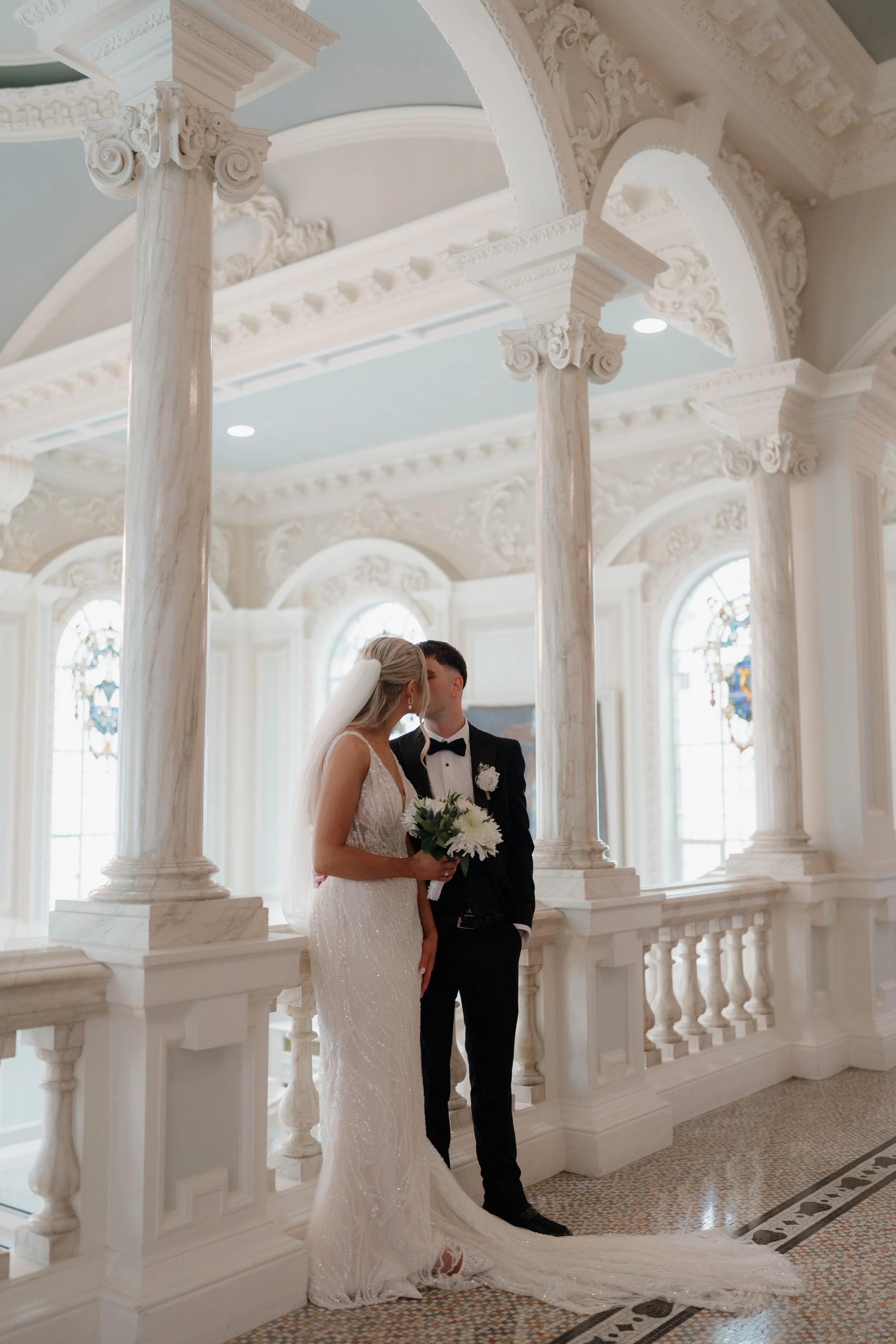 A bride and groom kissing in a church with ornate white columns and stained glass windows.