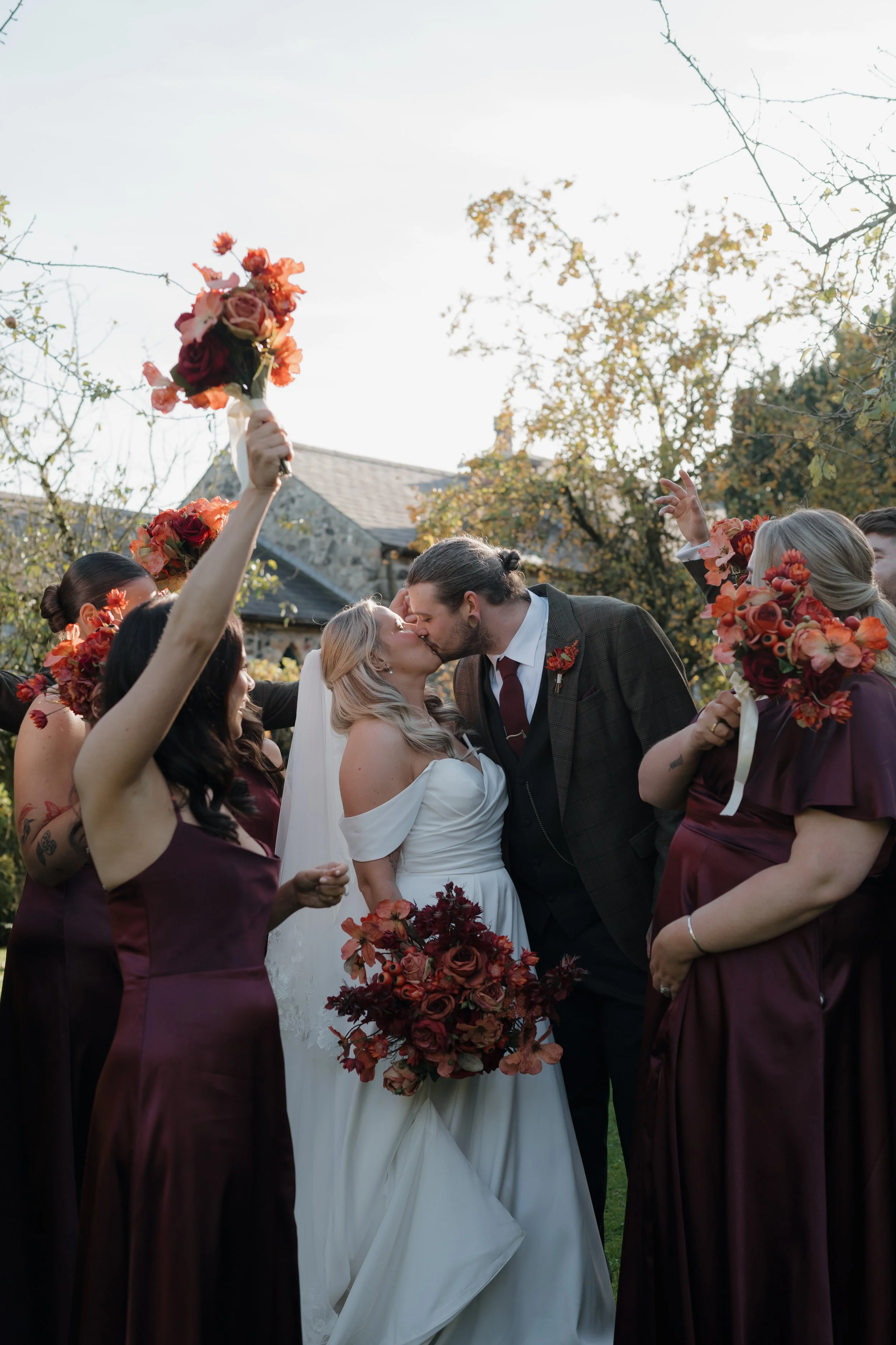 Bride and groom kiss during their outdoor wedding ceremony, surrounded by bridesmaids in burgundy dresses holding bouquets of red and orange flowers, with trees and a house in the background.