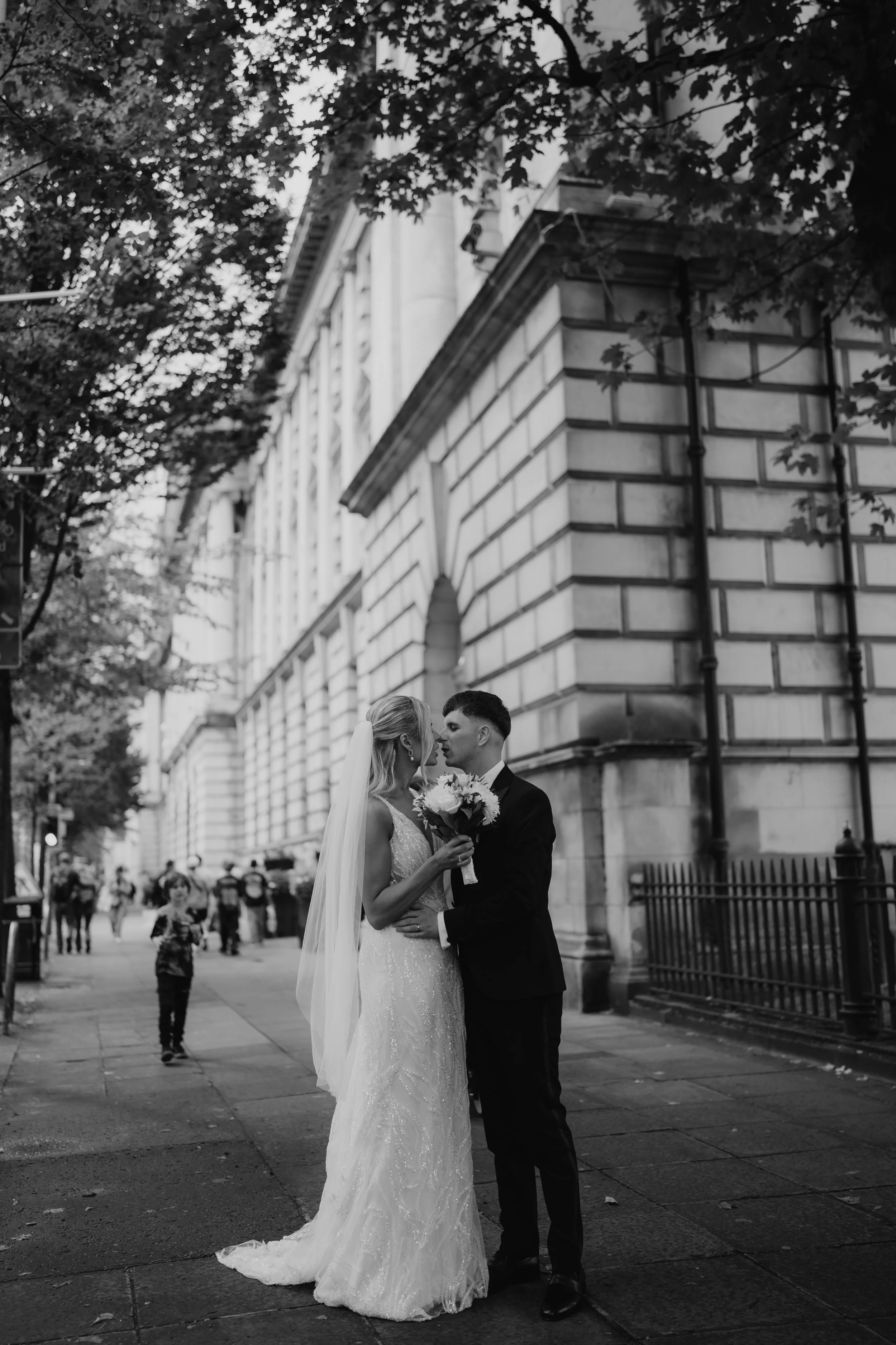 A bride and groom share a tender moment on a city street, with the bride holding a bouquet of flowers, surrounded by trees and a historic building in the background.