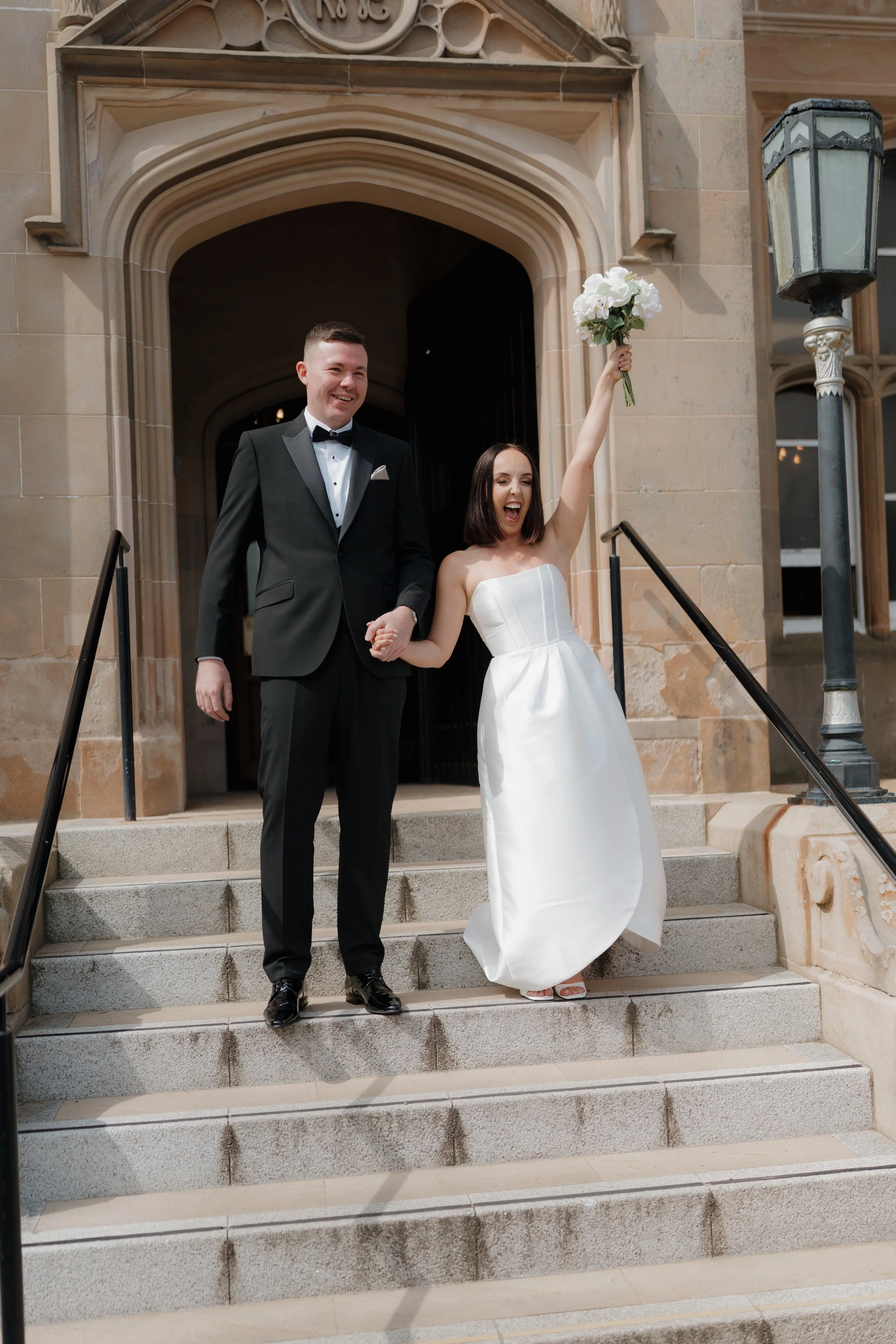 A newlywed couple standing on steps outside a church. The bride, in a strapless white wedding gown, is holding a bouquet of white flowers and raising it in celebration. The groom, in a black tuxedo with a bow tie, is smiling and holding her hand.