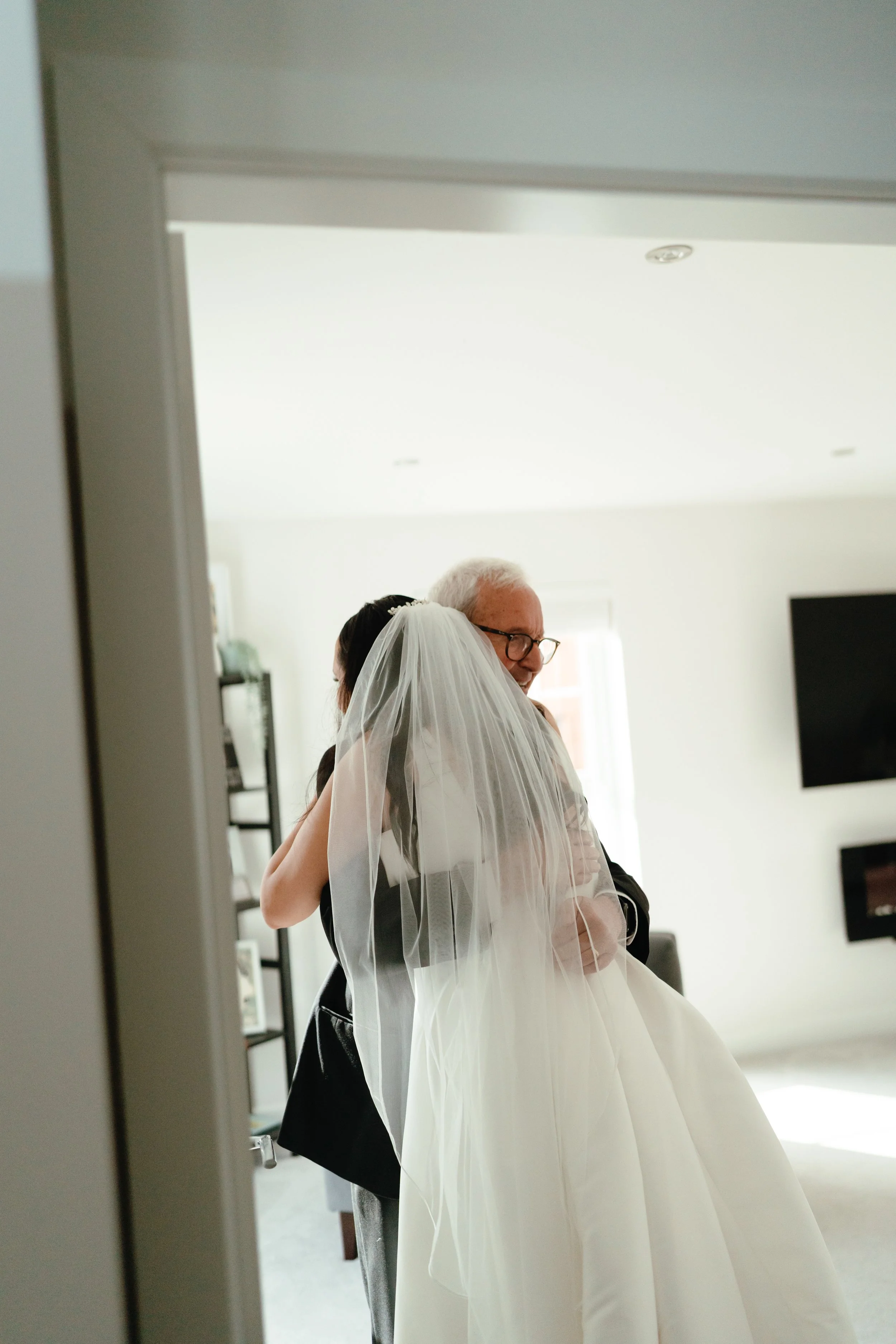 A bride in a white wedding dress and veil hugging an older man, likely her father, in a bright room, seen through a doorway.