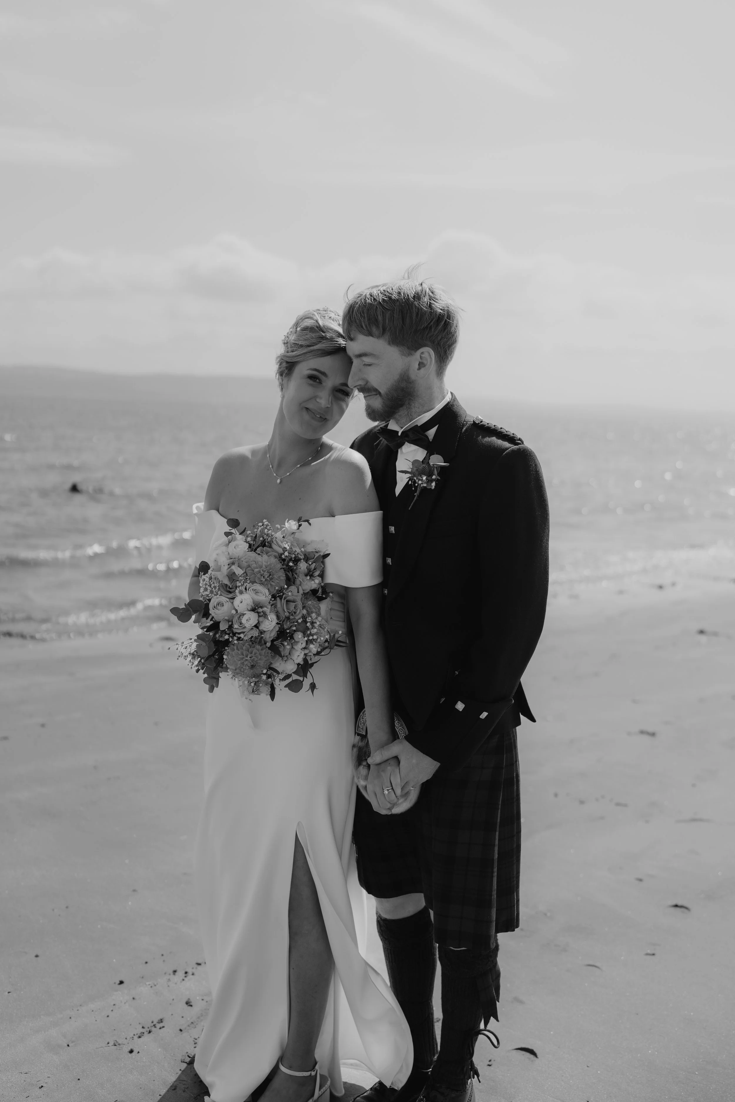 A black and white photo of a bride and groom on the beach, holding hands and smiling.