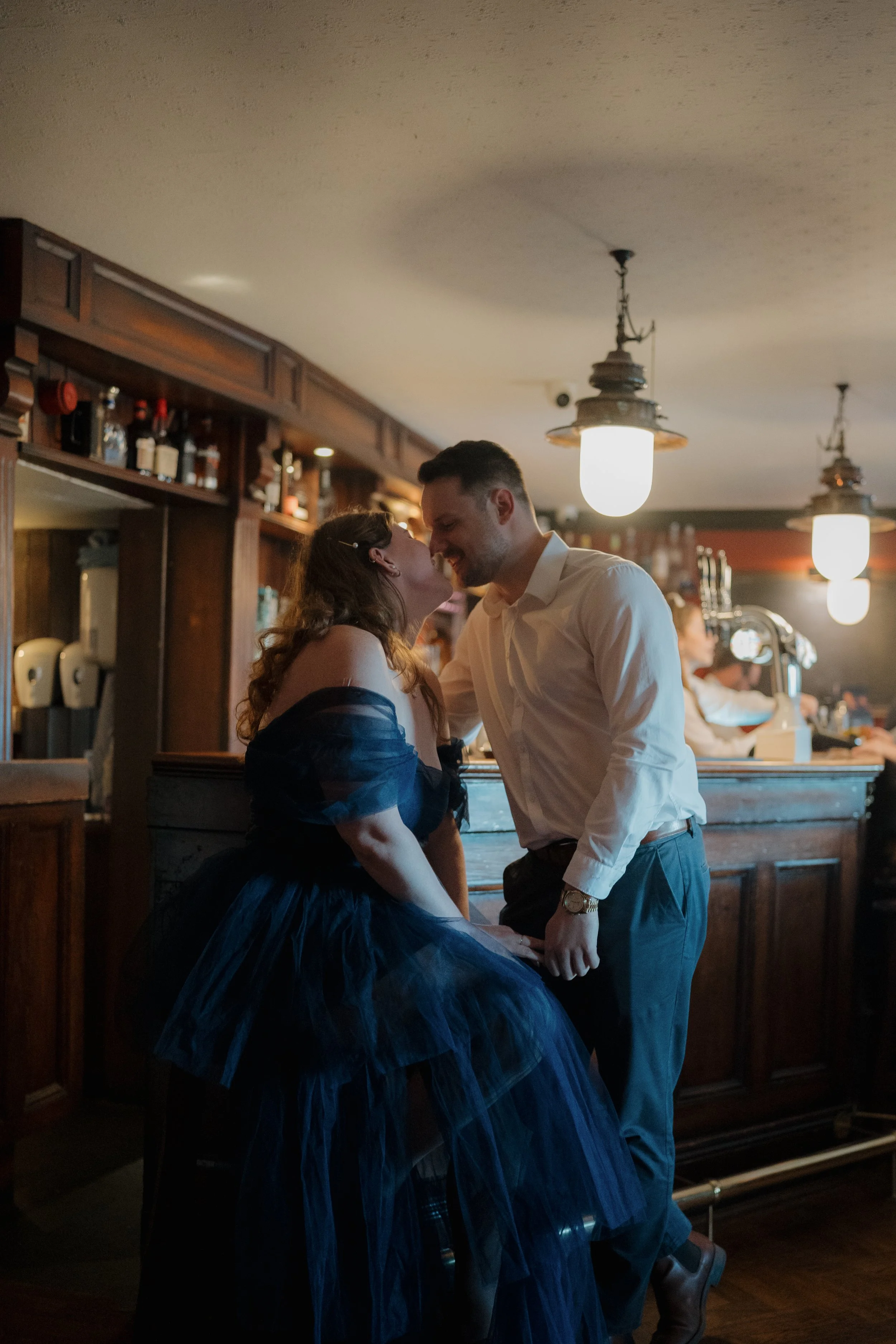 A couple sharing a romantic moment at a bar, with the woman in a dark dress and the man in a white shirt, smiling and leaning close to each other.