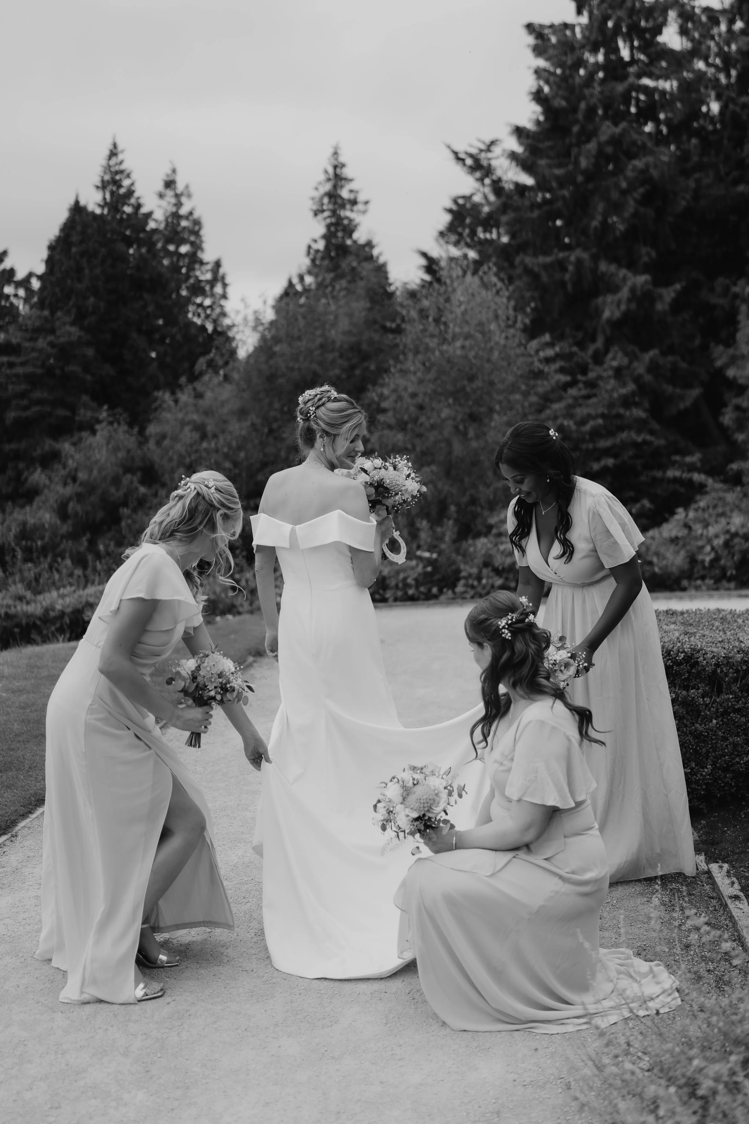 A bride in a wedding dress holding a bouquet, surrounded by four bridesmaids in light-colored dresses, outdoors with trees in the background at Belfast Castle.