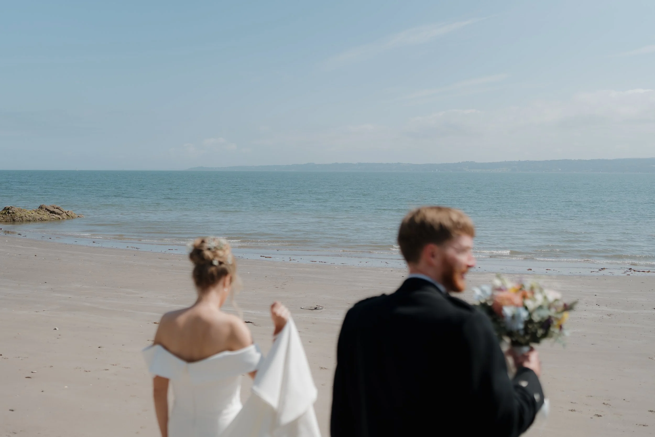 A bride and groom on a beach, with the bride holding part of her dress and the groom holding a bouquet, under a partly cloudy sky.