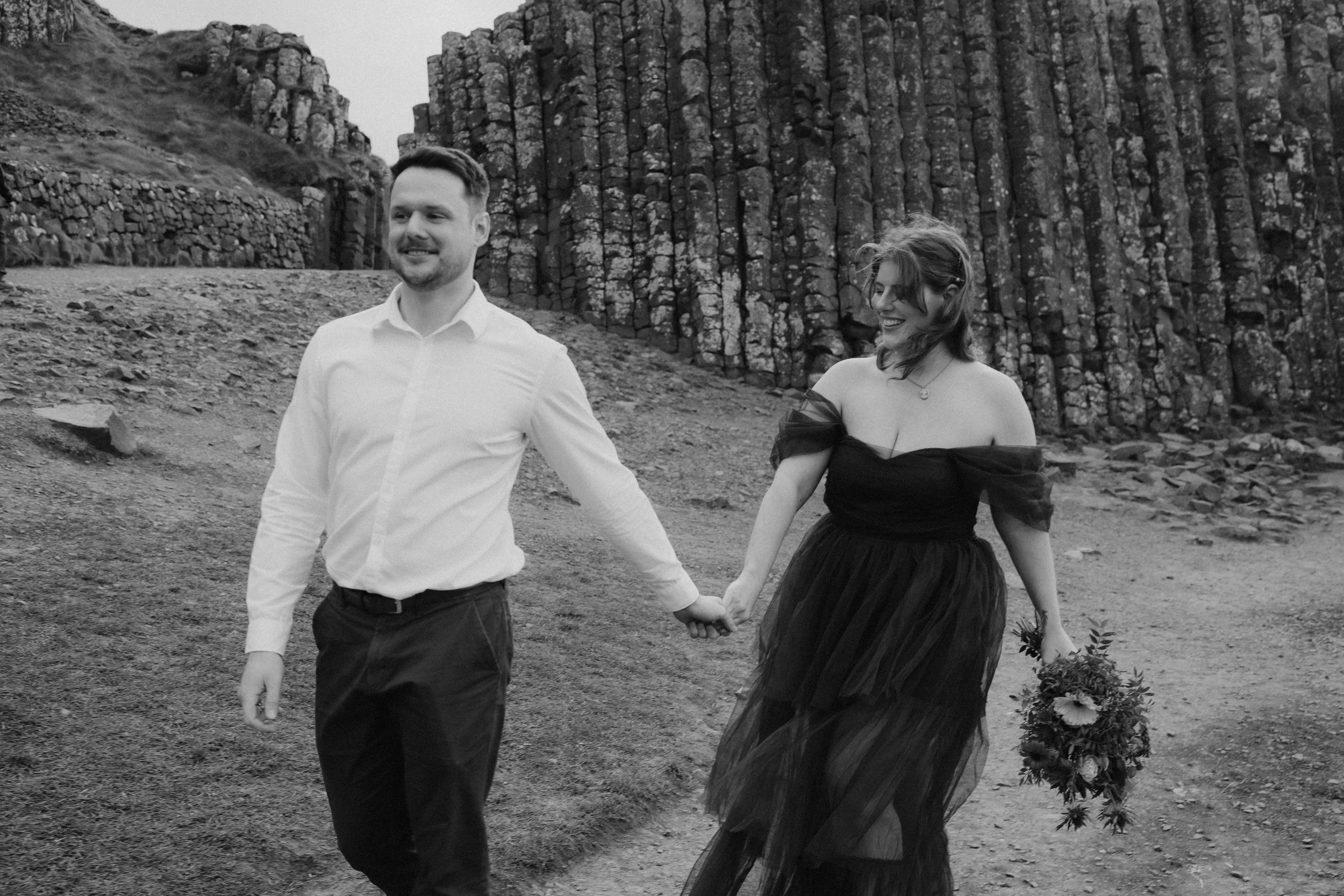 A happy couple holding hands and walking outdoors near large rocks, with the woman holding a bouquet of flowers, in black and white at the Giants Causeway.