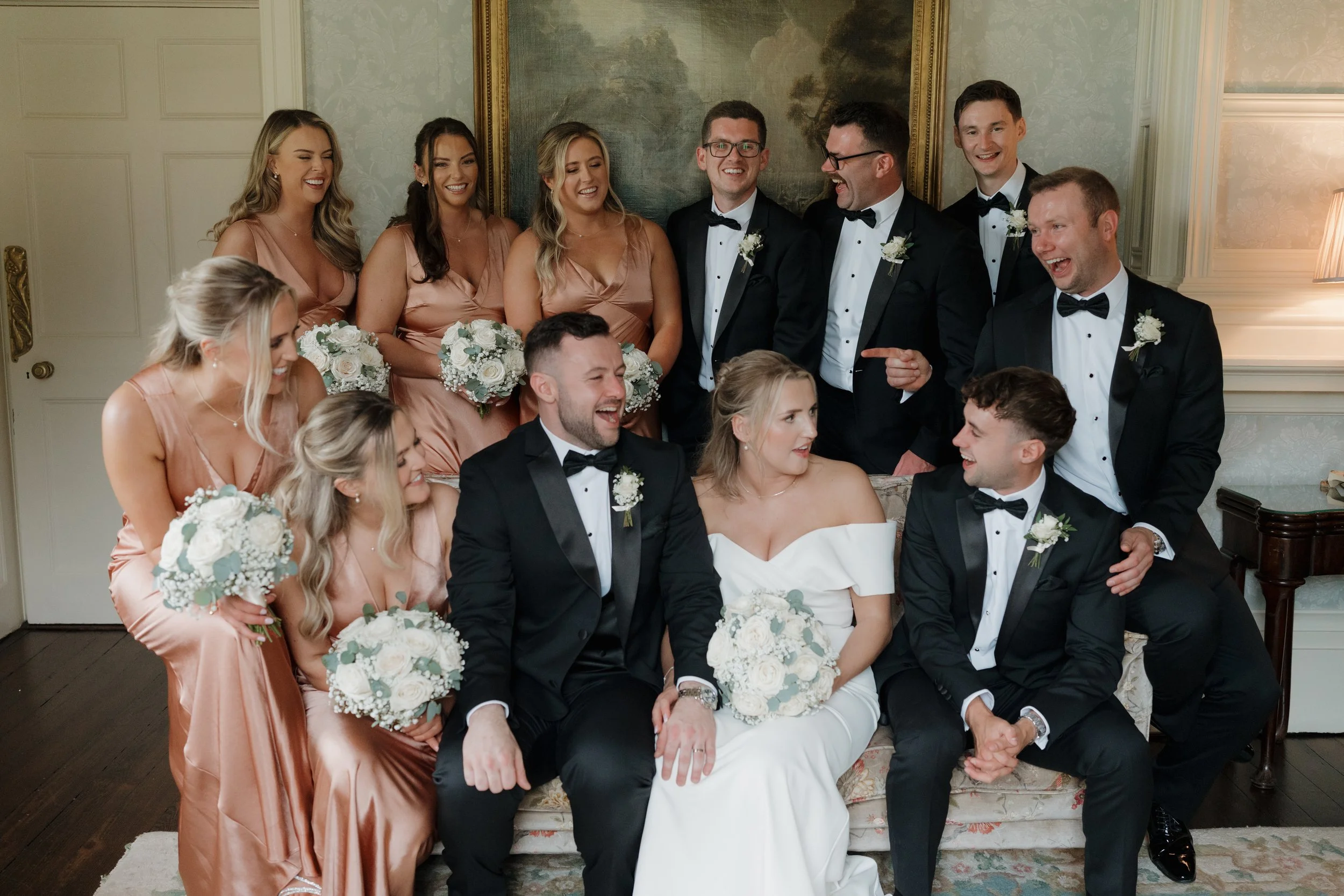 A wedding party with the bride and groom sitting on a sofa, surrounded by bridesmaids in peach dresses holding bouquets and groomsmen in black tuxedos with bow ties, all smiling and laughing indoors.