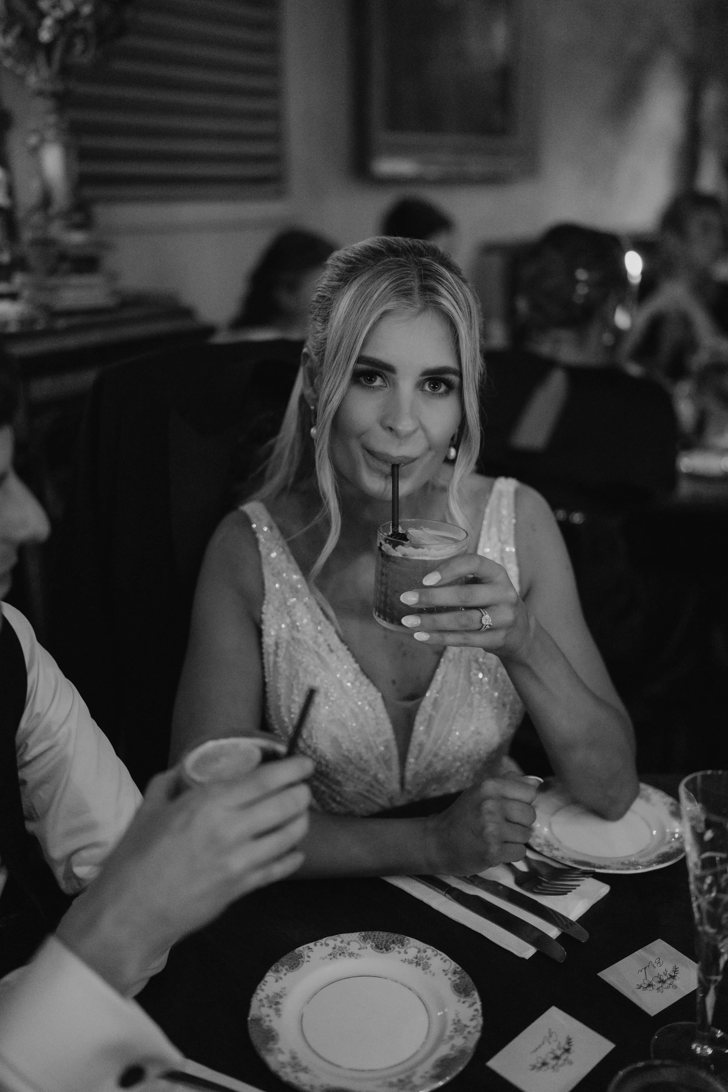 A woman in a sleeveless, sparkly dress drinks from a glass with a straw at a dinner event.