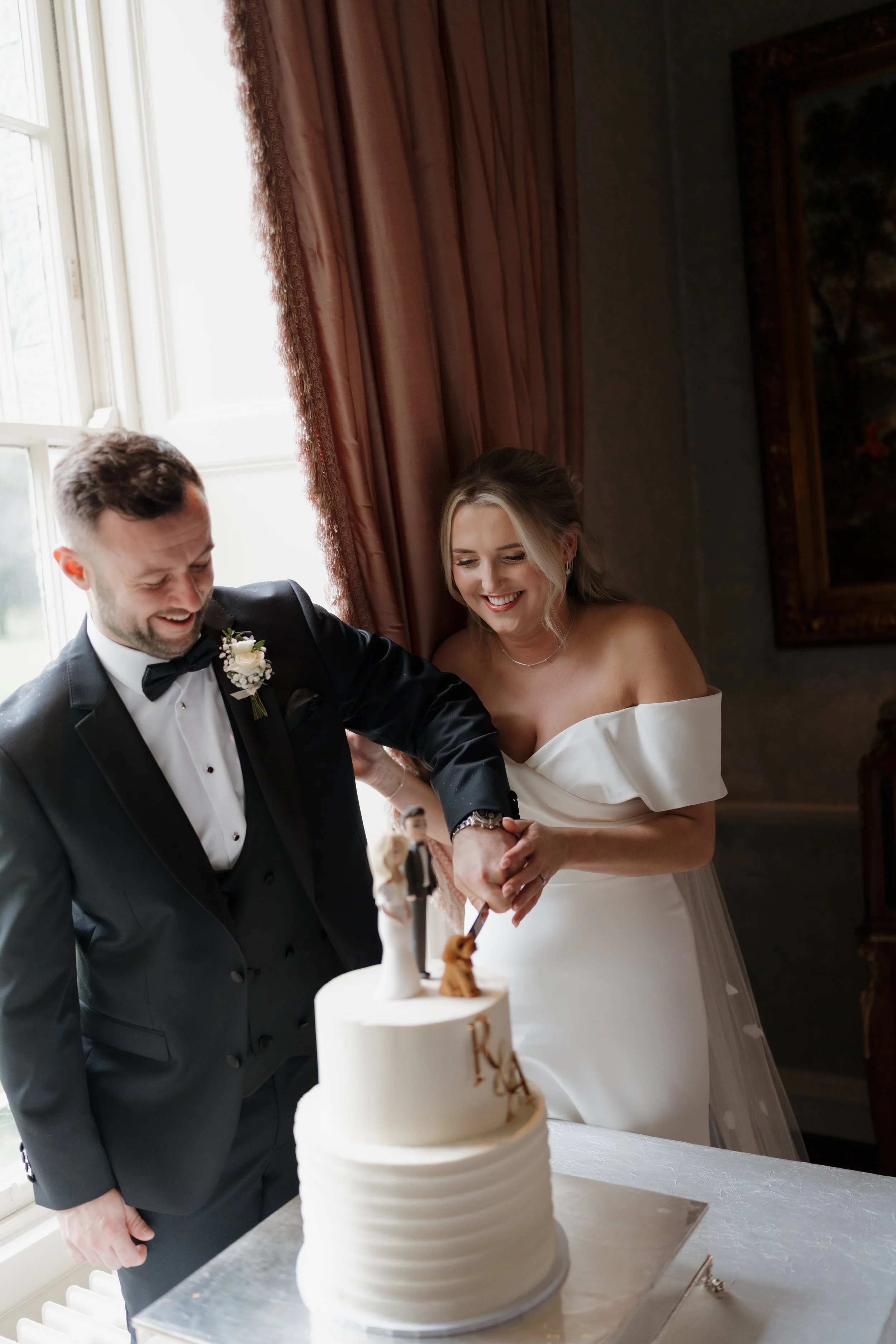 Bride and groom cutting a wedding cake together, both smiling, in a well-lit room with large window and curtains.
