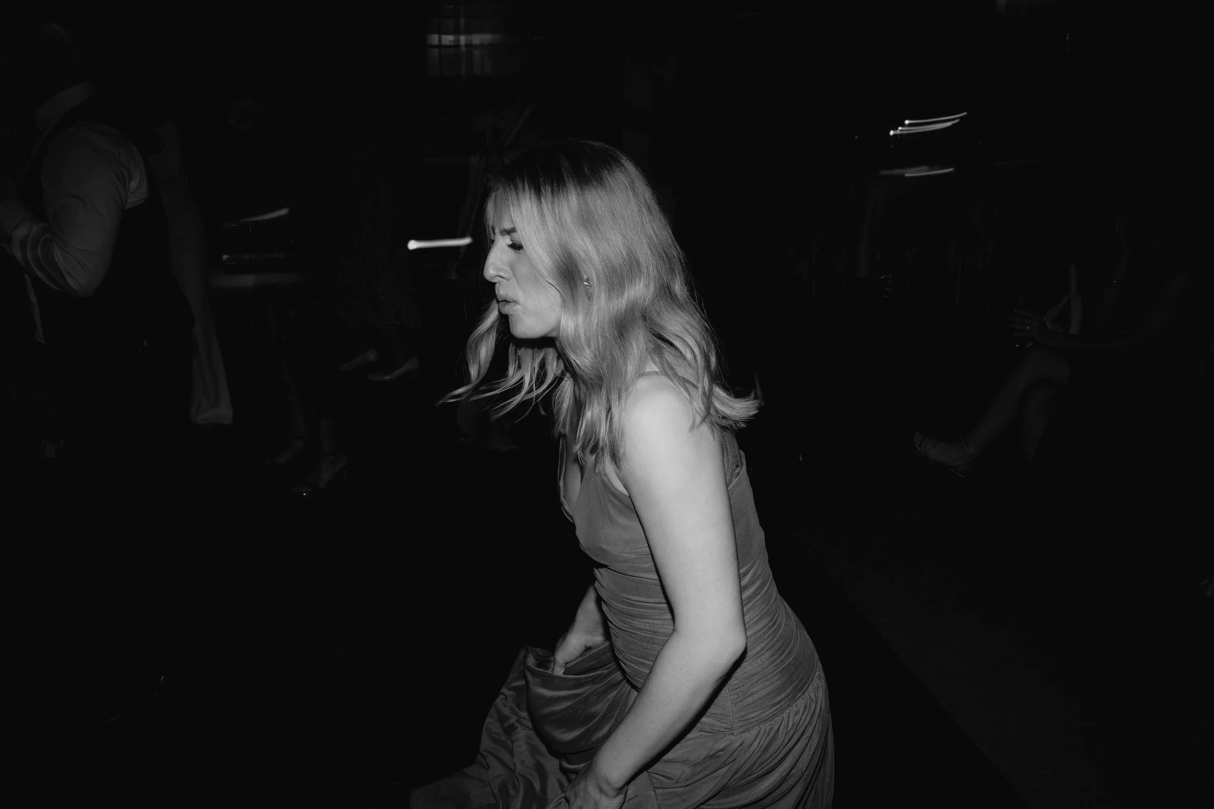 A woman with wavy blonde hair in a sleeveless dress is walking in a dark room.