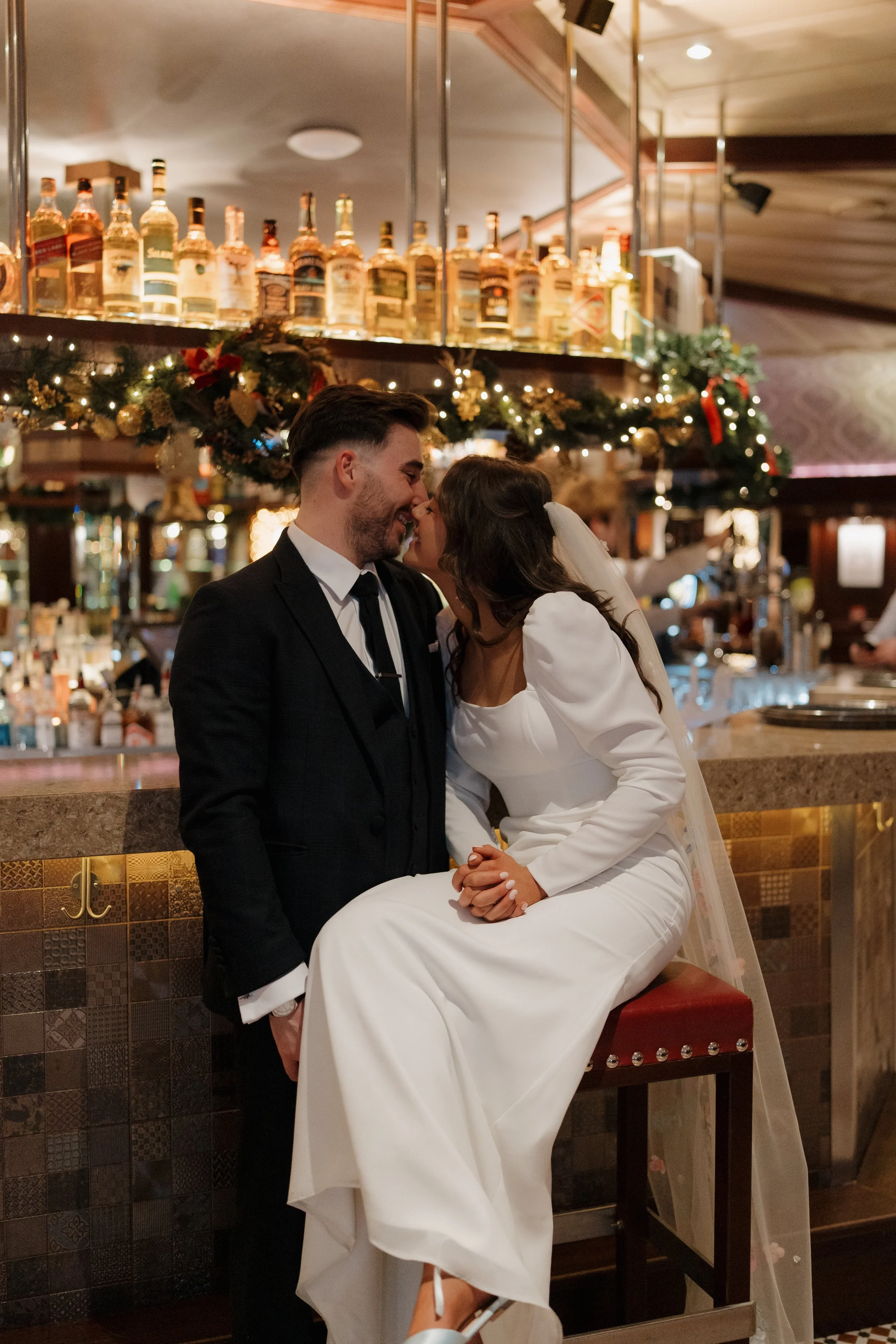 A newlywed couple sharing a kiss at a bar, with Christmas decorations and liquor bottles in the background.