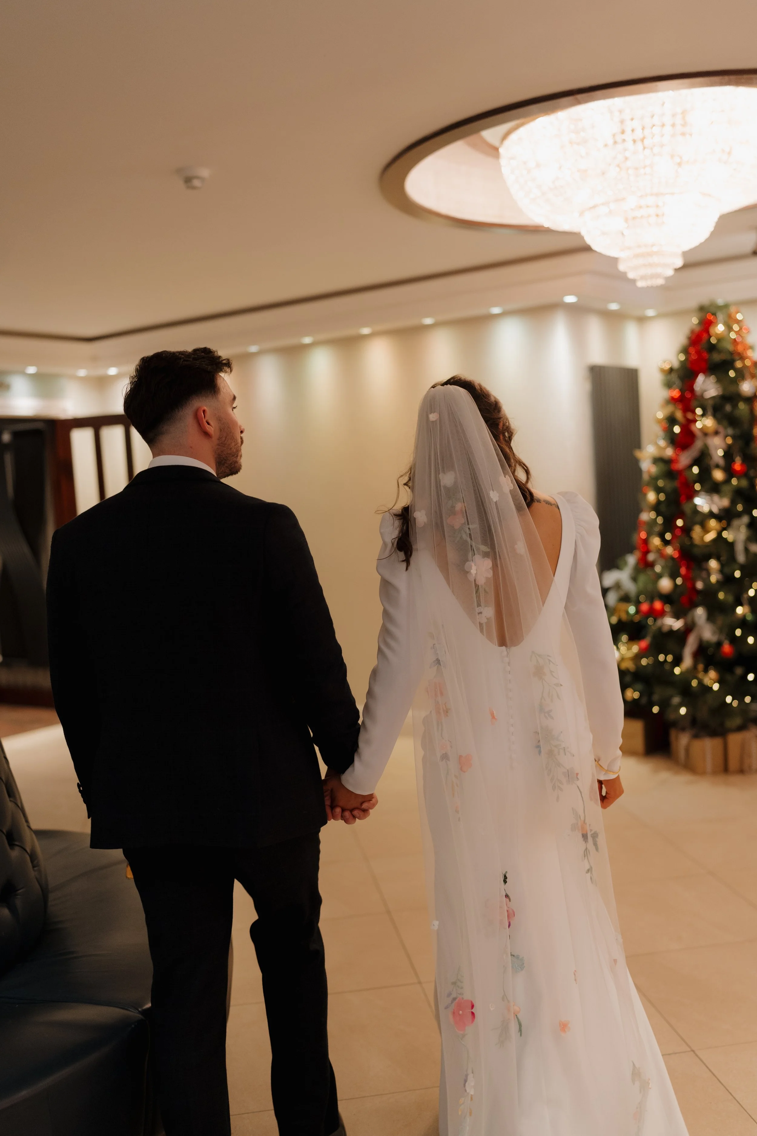 A bride and groom hold hands during their wedding ceremony in an elegant indoor setting with a Christmas tree decorated with red and gold ornaments in the background at Harveys Point, Donegal.