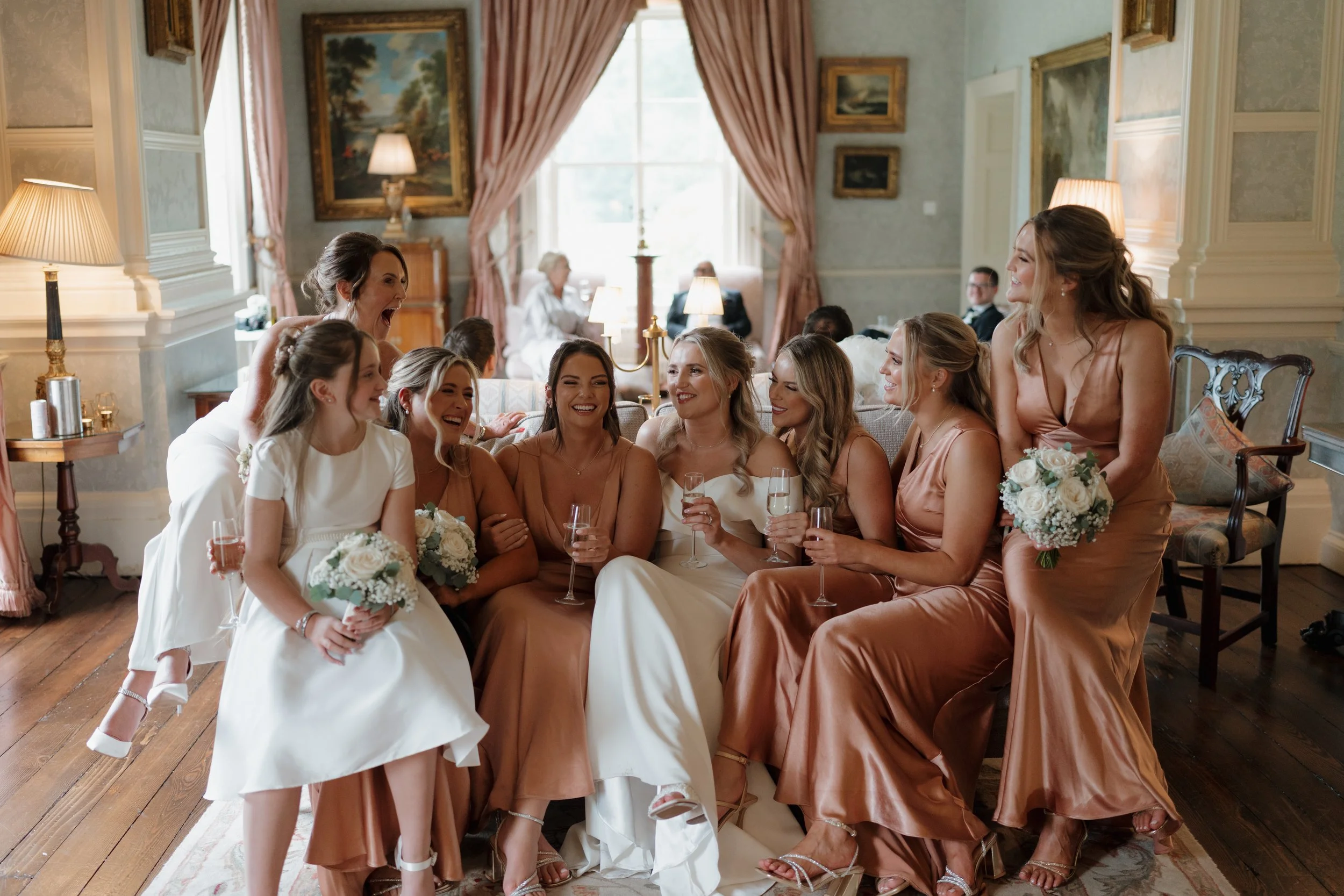 A bride and bridesmaids relaxing and celebrating, holding glass of champagne, in a vintage-style room with artwork and curtains.