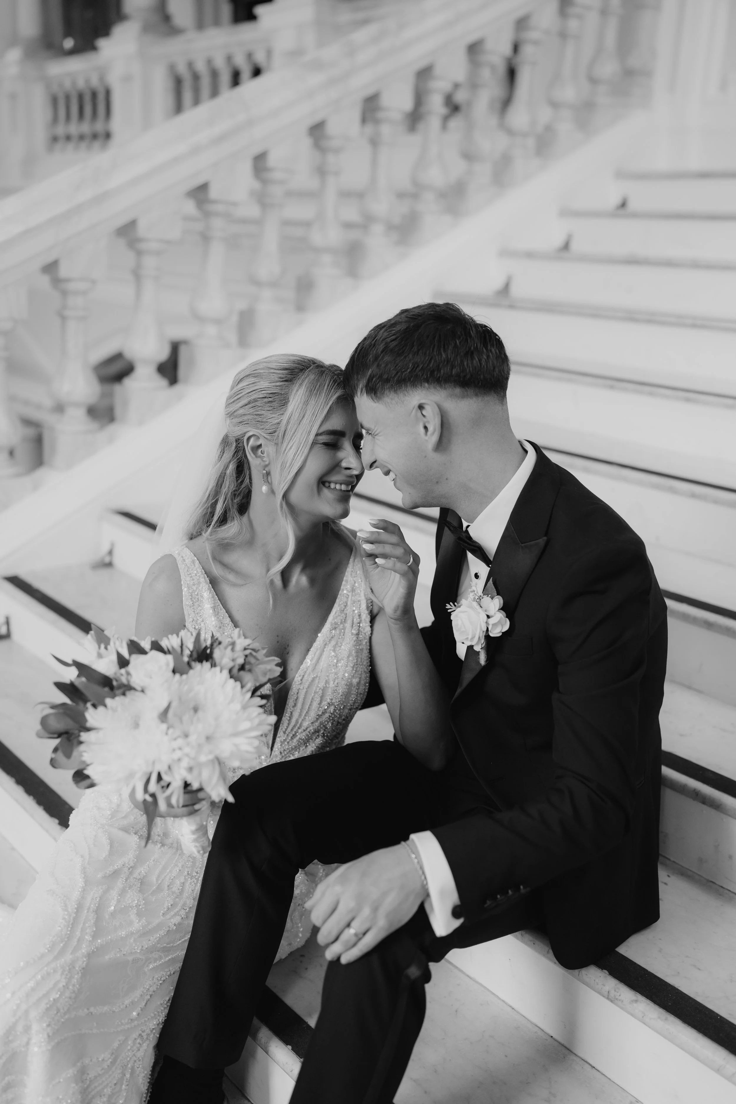 A black-and-white photo of a bride and groom sitting on stairs, smiling and touching foreheads, with the bride holding a bouquet of flowers.