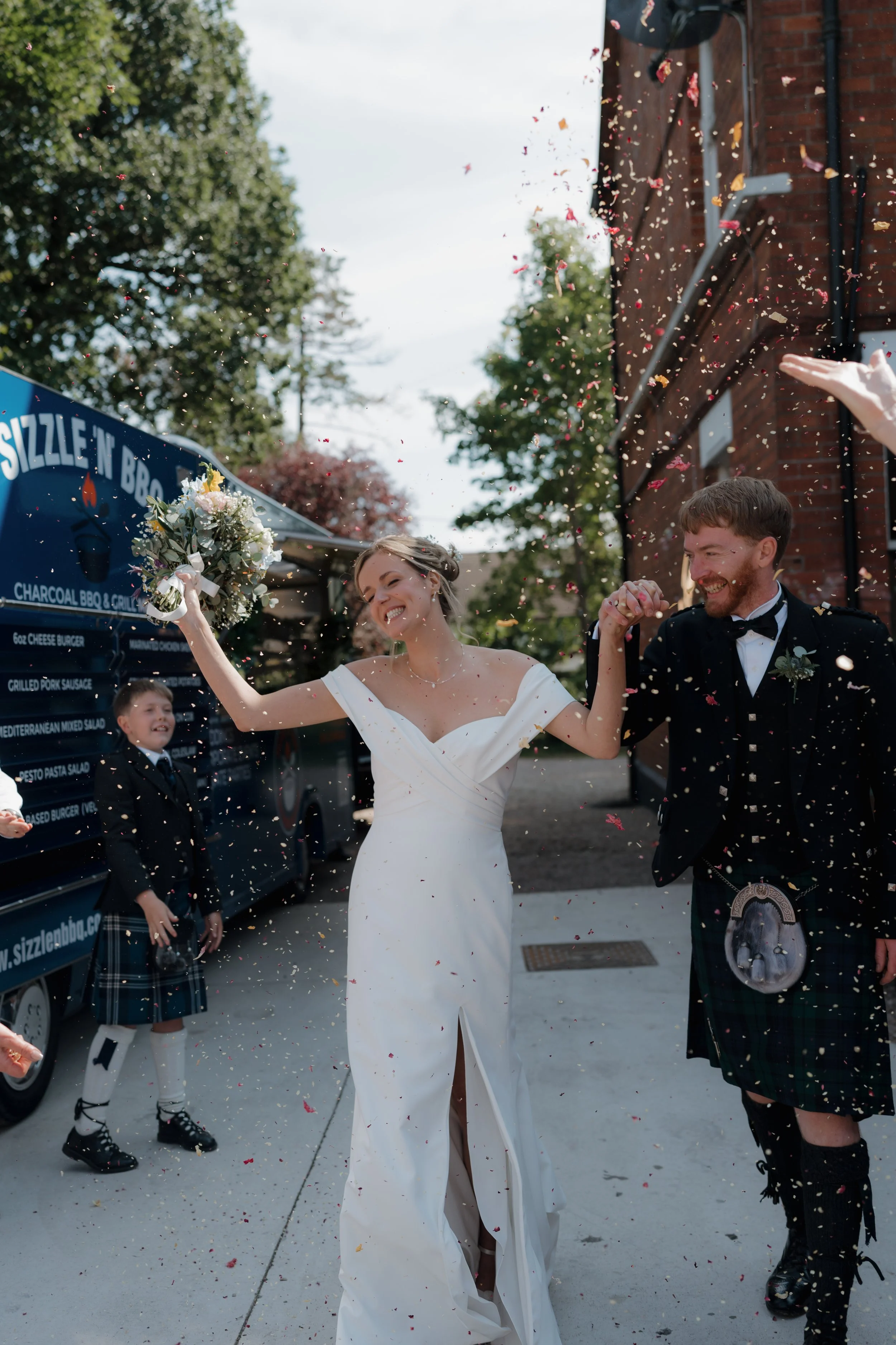 A bride and groom celebrating outdoors, holding hands, with confetti falling around them, and children watching in the background.