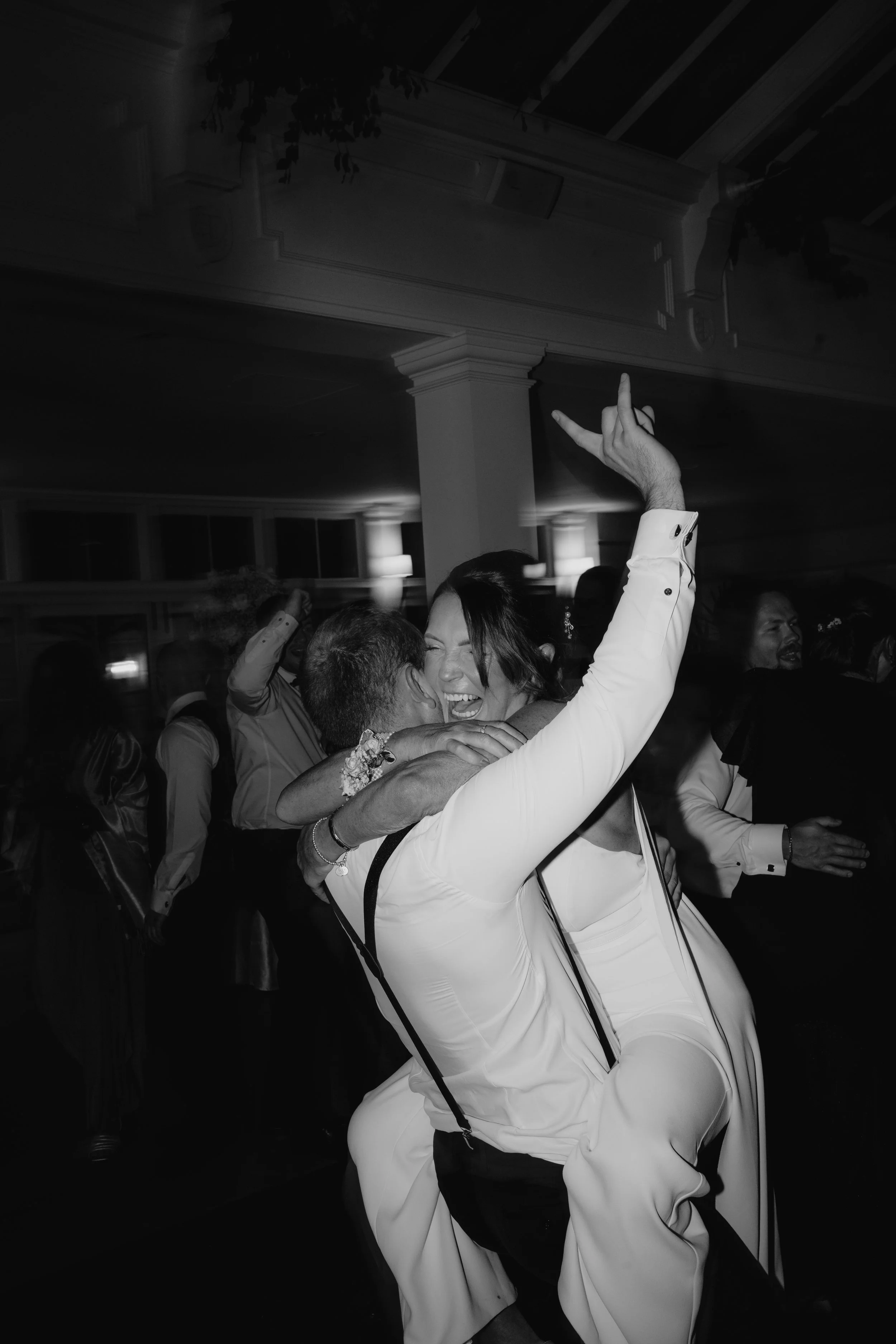 People dancing and celebrating at a wedding reception, with a woman and a man hugging, and a woman in a white dress raising her arm in joy.