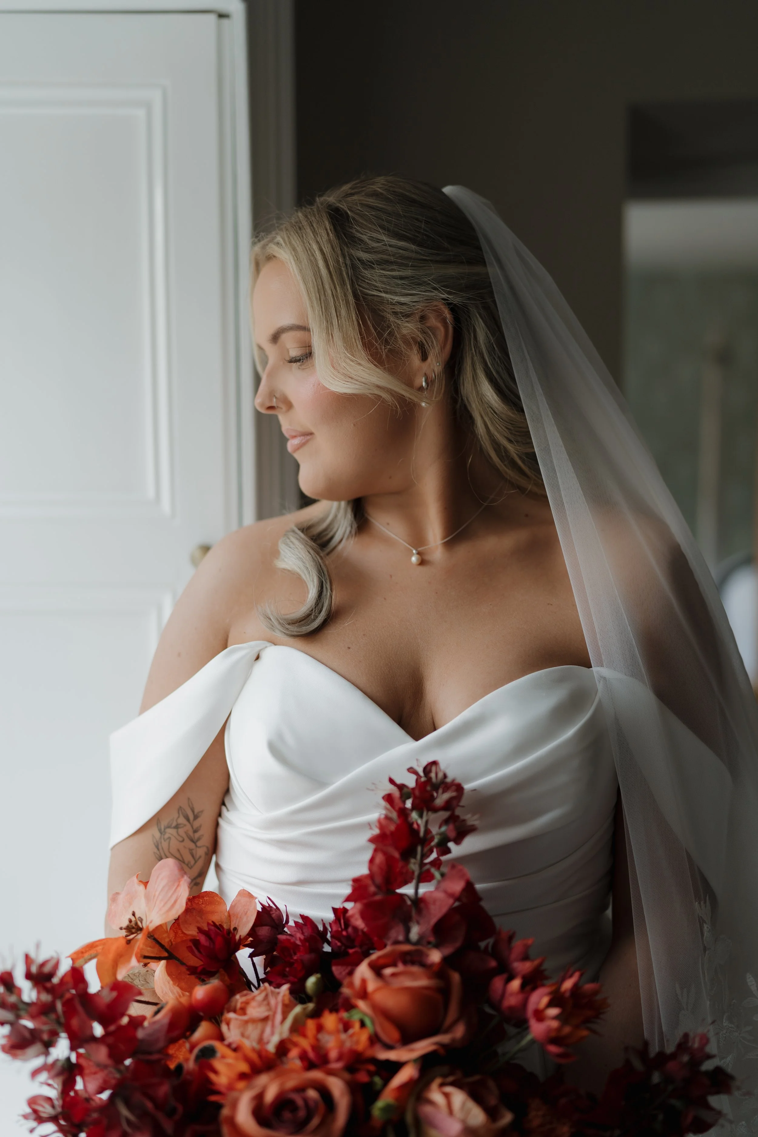 Bride in white wedding dress holding a bouquet of red, orange, and pink flowers, wearing a veil, looking down, standing near a window.