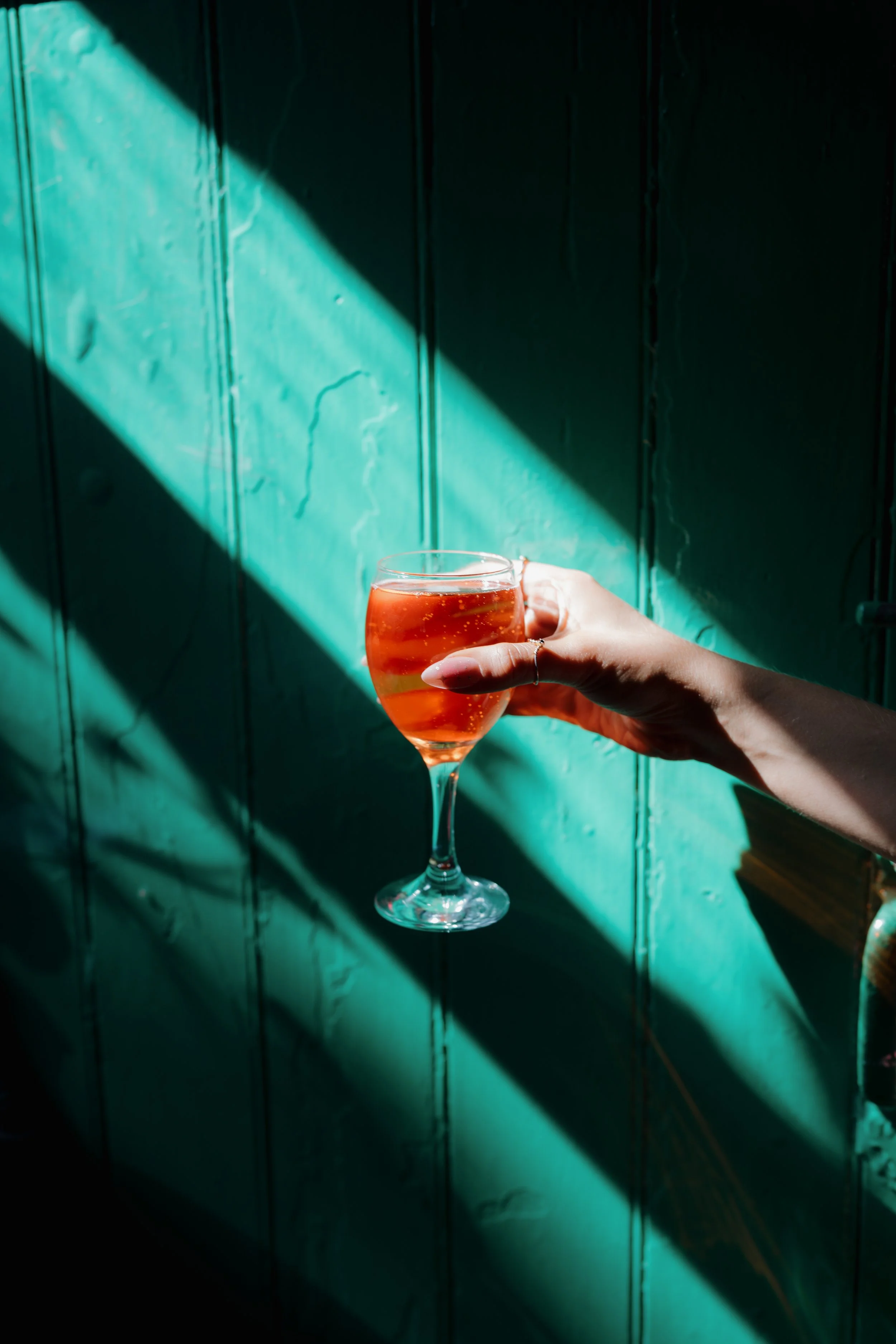 Person holding a glass of pinkish-orange beverage against a teal wooden wall with sunlight and shadow patterns.