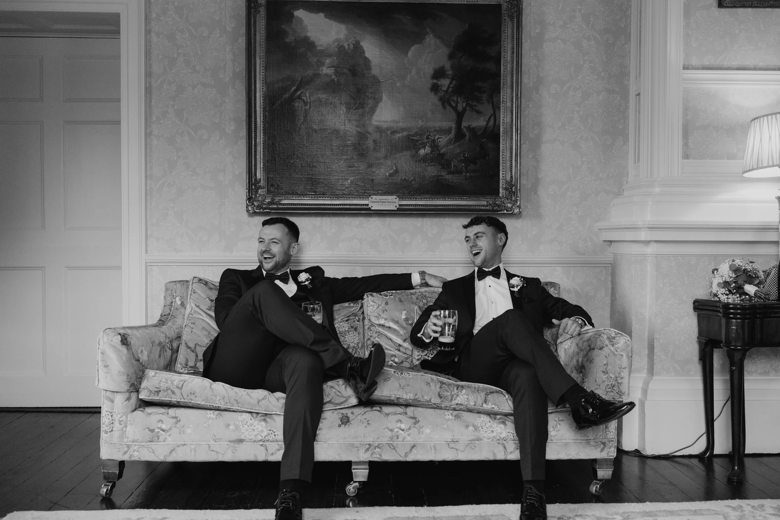 Two men in tuxedos sit on a floral upholstered couch, laughing and holding glasses of beer, in a room with ornate wallpaper and a large landscape painting on the wall behind them.