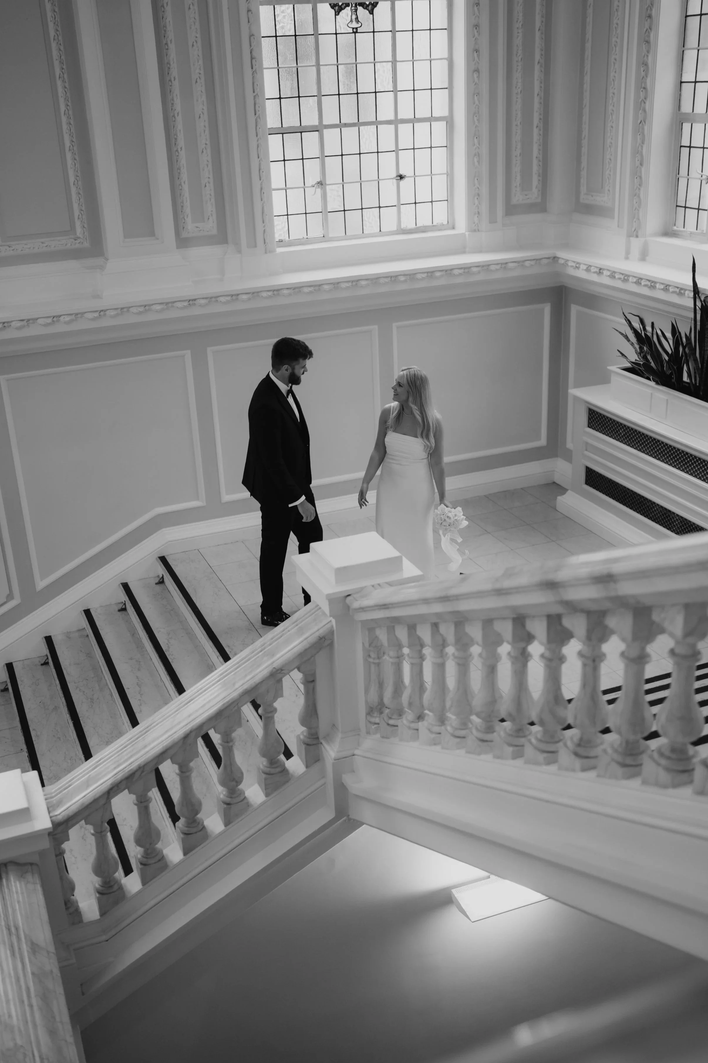 A black and white photo of a bride and groom standing on a staircase inside a bright, ornate room with large windows in Belfast City Hall, engaged in a moment of connection.