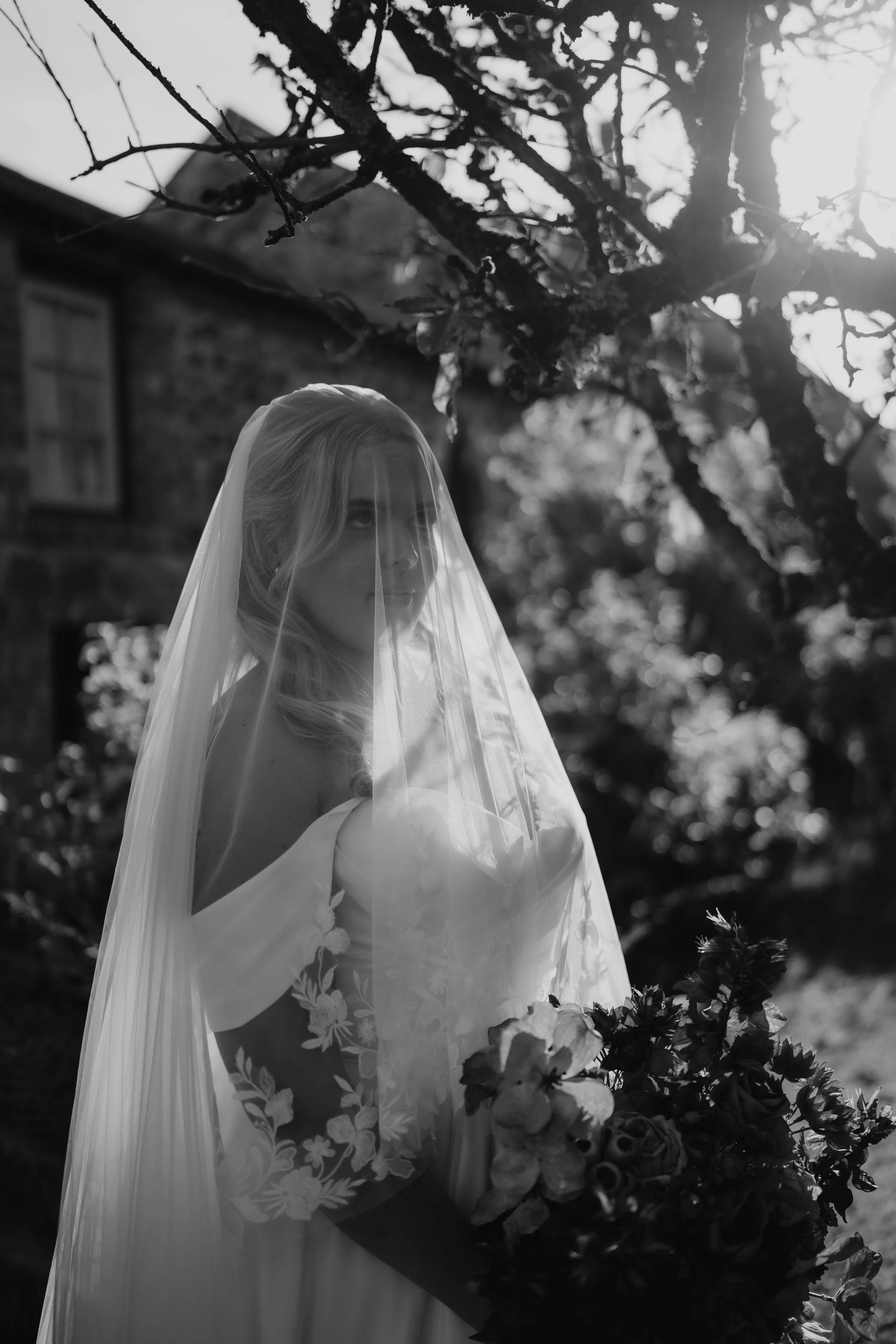 Black and white photo of a bride holding a bouquet of flowers, standing outdoors under a tree with sunlight shining through the branches.