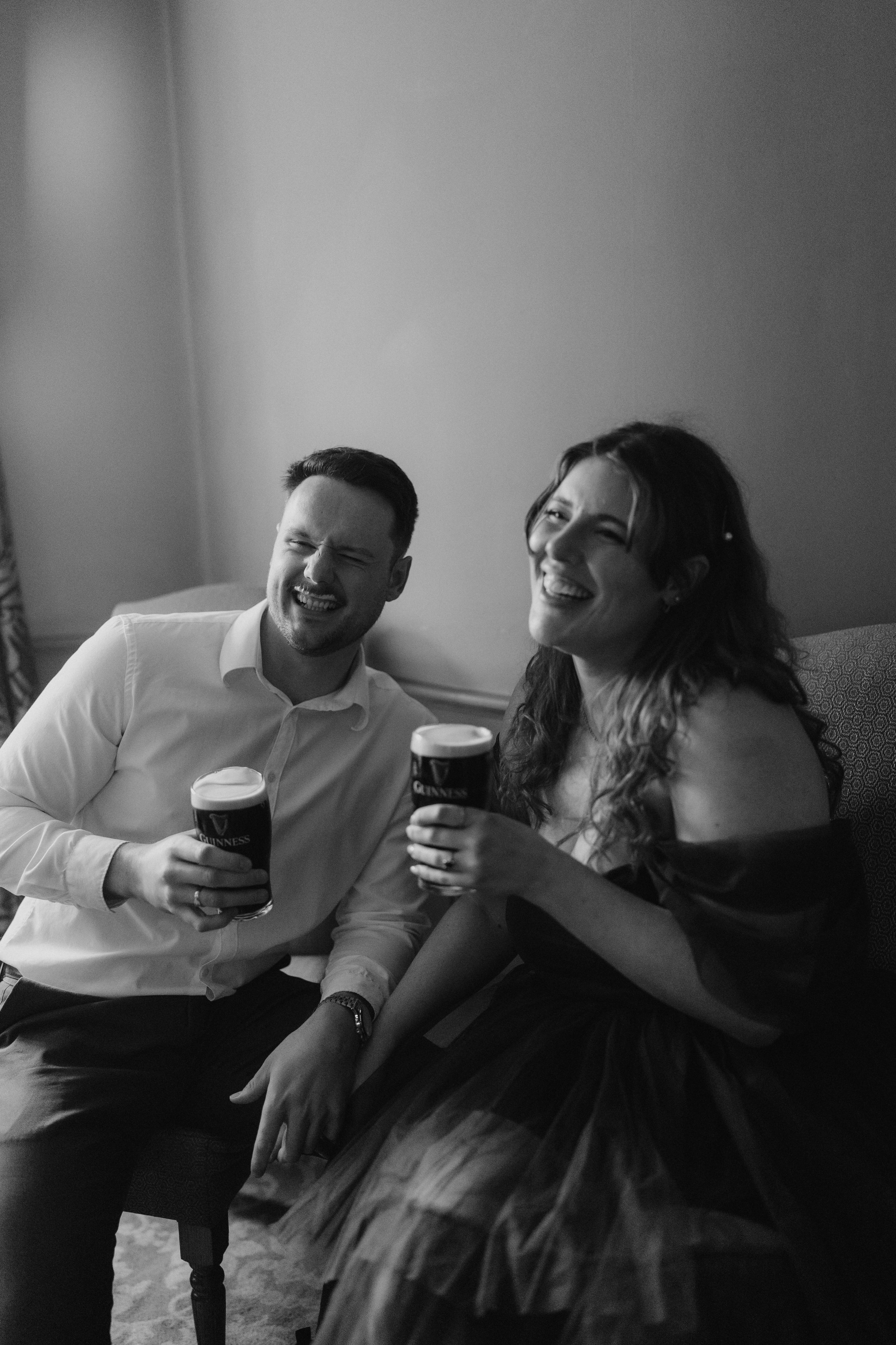 A black and white photo of a man and woman sitting on a sofa, both holding pints of Guinness beer and laughing.