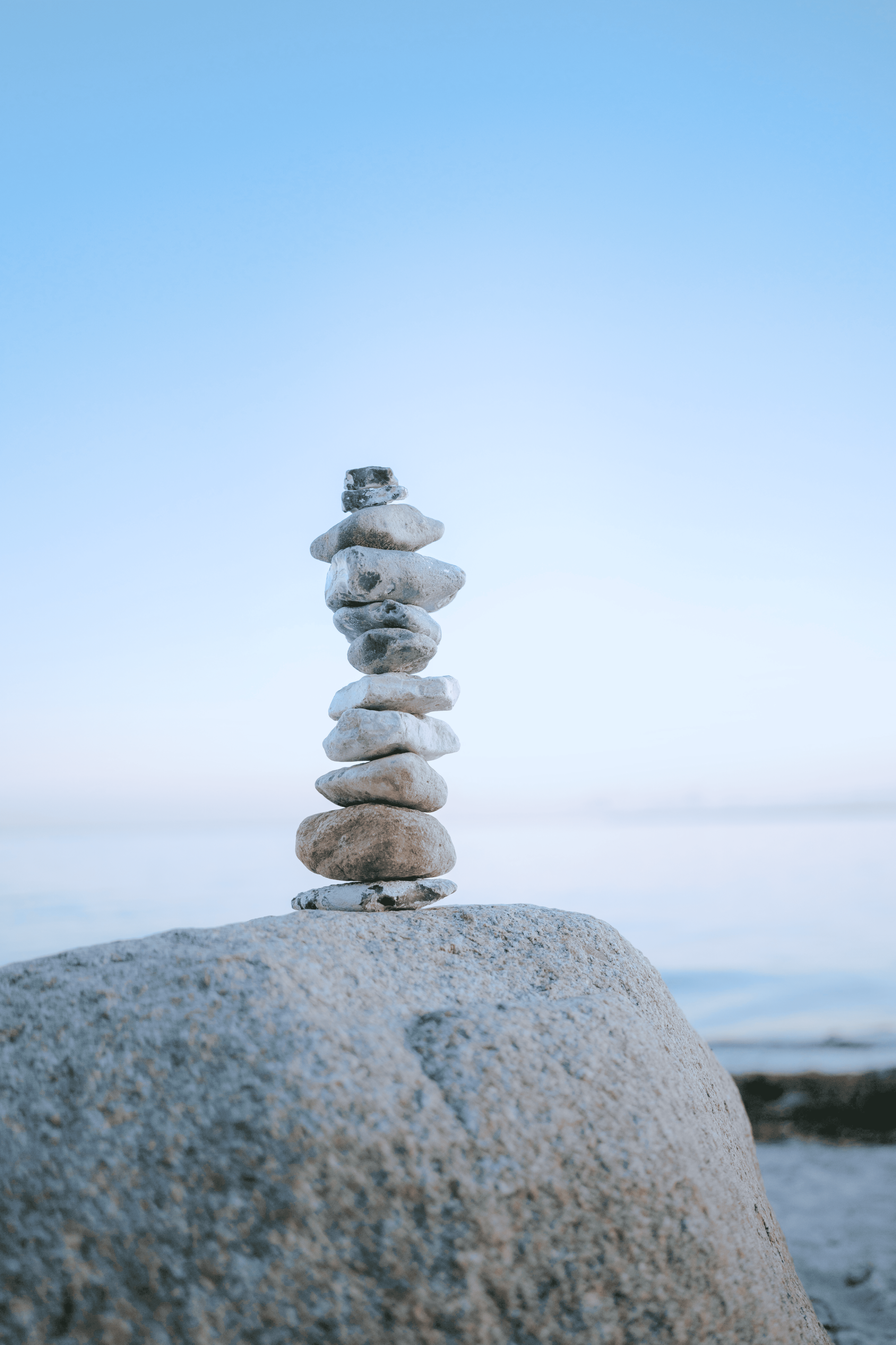 A stack of nine smooth stones balanced on a large rock by the water on a clear day.