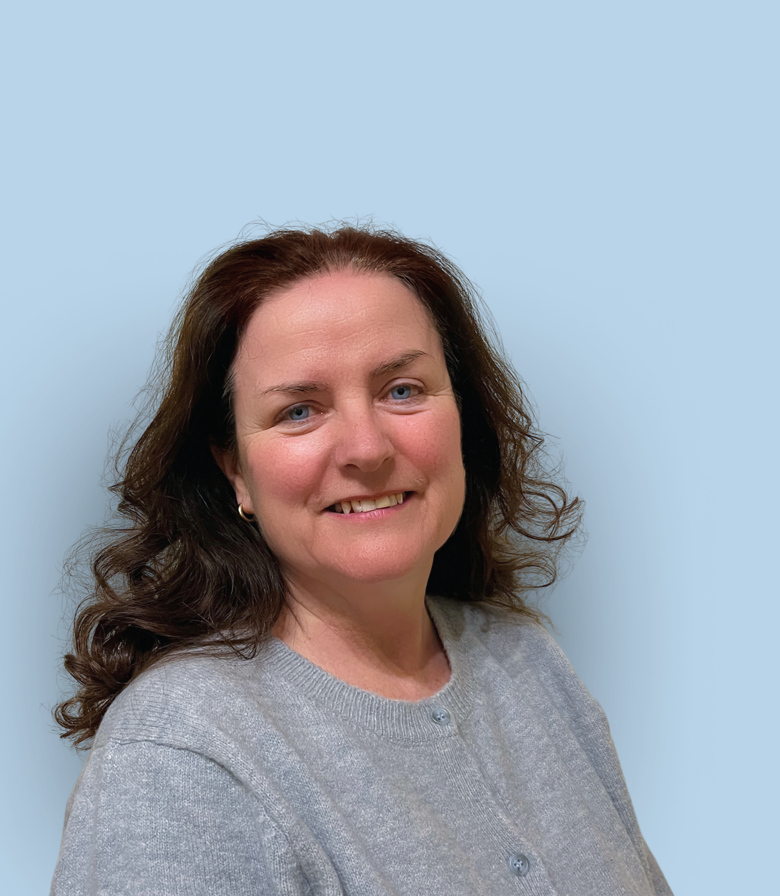 A portrait of a woman with long brown curly hair, blue eyes, and a friendly smile, wearing a light gray sweater, standing against a light blue background.